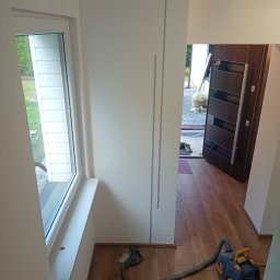 Krys-Bud usługi ogólnobudowlane Krystian Szweda - Jasne wnętrze po remoncie: białe ściany z subtelnymi szarymi akcentami, drewnopodobna podłoga i okno z widokiem na zieleń. Odkurzacz na podłodze sugeruje prace porządkowe.