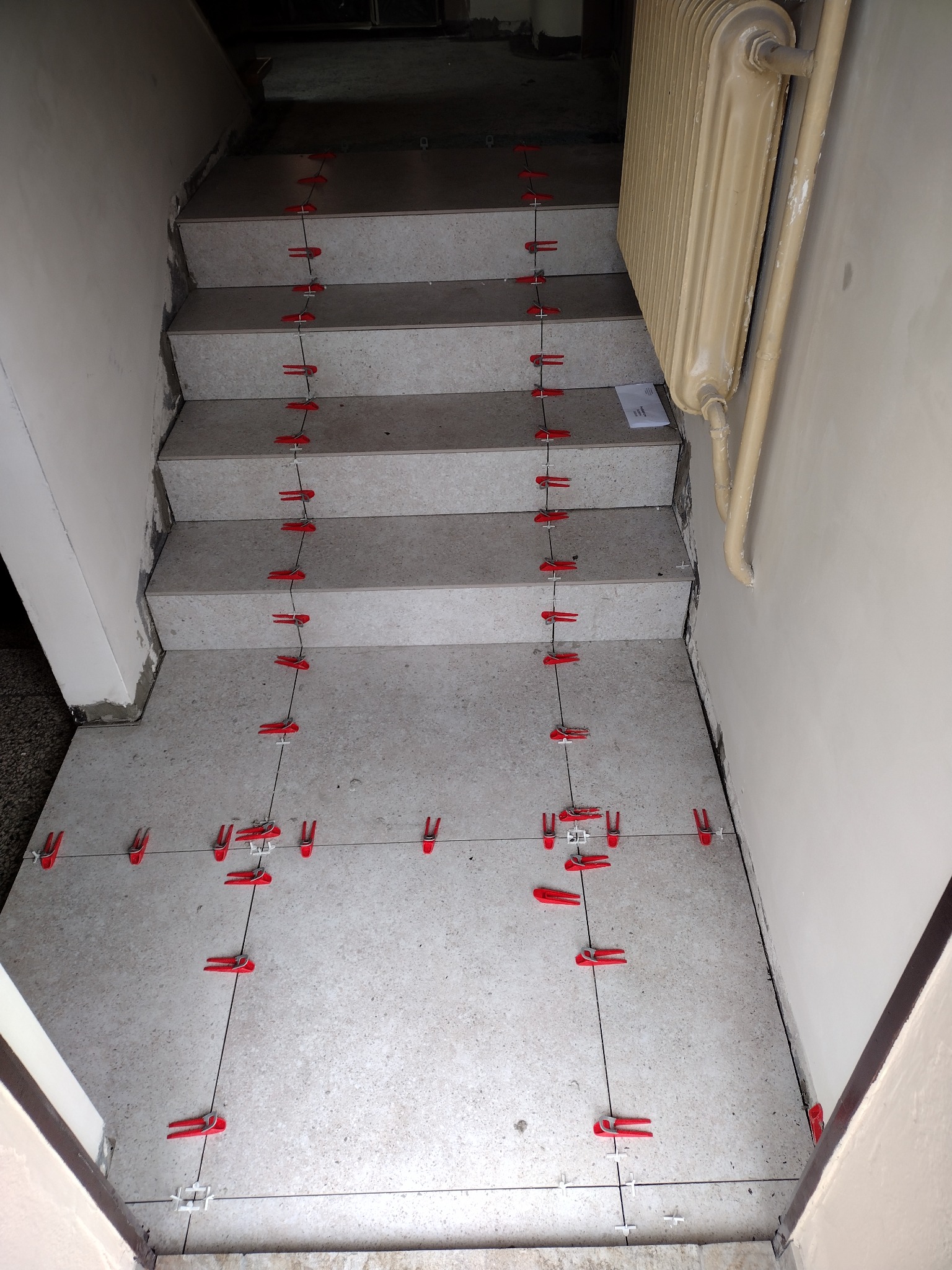 Klatka schodowa w trakcie układania płytek, widoczne czerwone klipsy poziomujące. Na ścianie stary grzejnik. Ujęcie z góry.