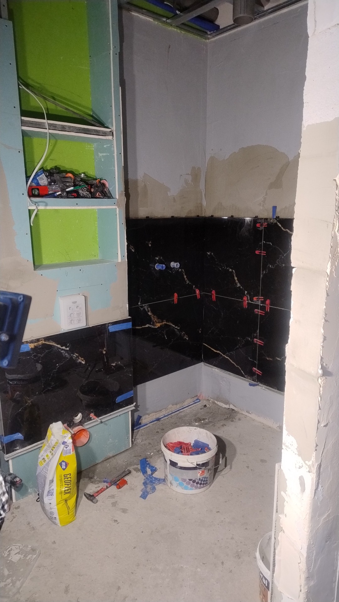 Częściowo wykończona łazienka z czarnymi płytkami imitującymi marmur, narzędzia glazurnicze i worek zaprawy widoczne na podłodze. Elementy zabudowy z płyt gipsowych.