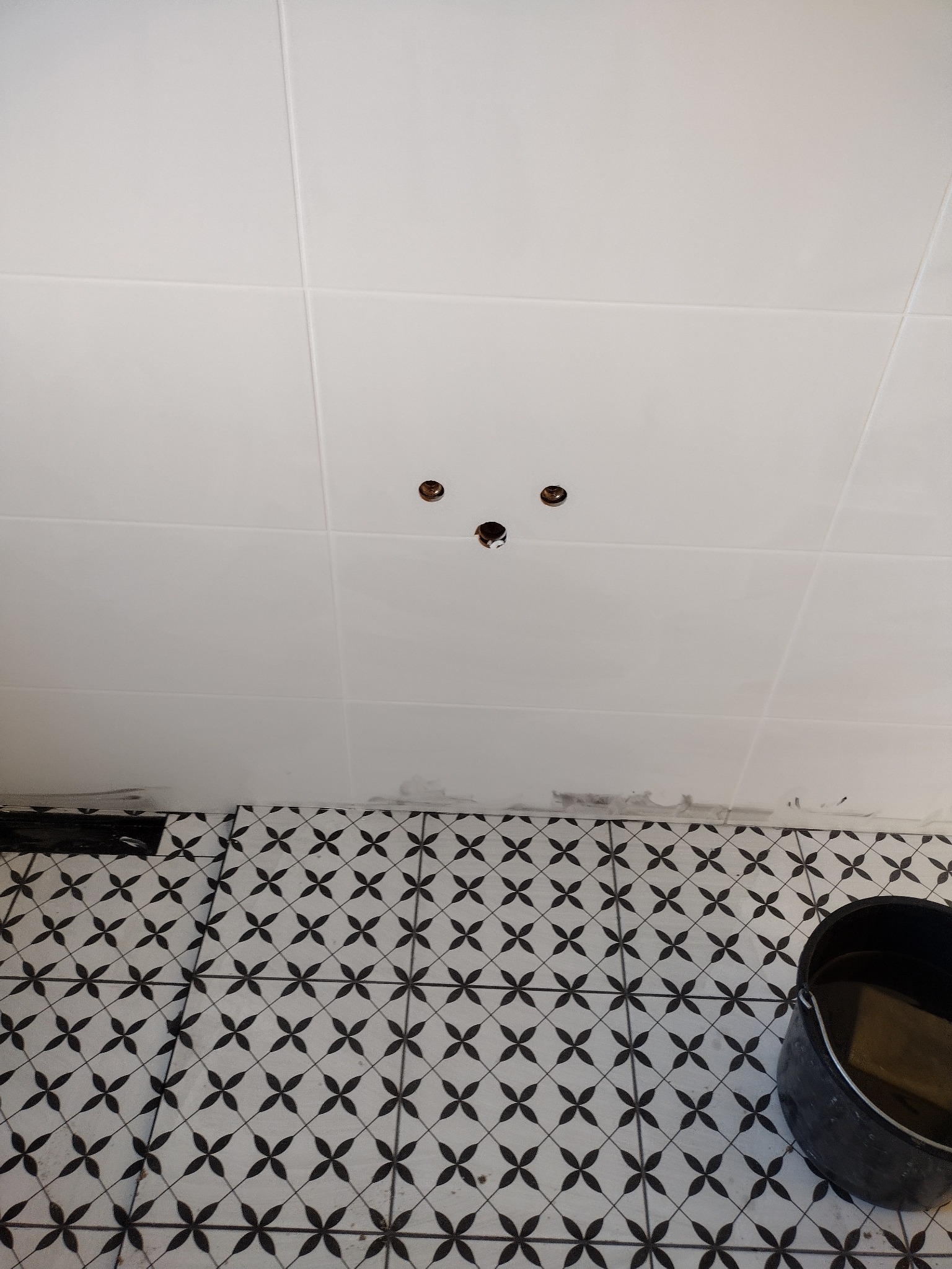 Białe płytki ścienne z otworami i wzorzysta podłoga w romby. Czarny kubeł w rogu. Fragment ułożonej podłogi. Prace wykończeniowe w łazience.