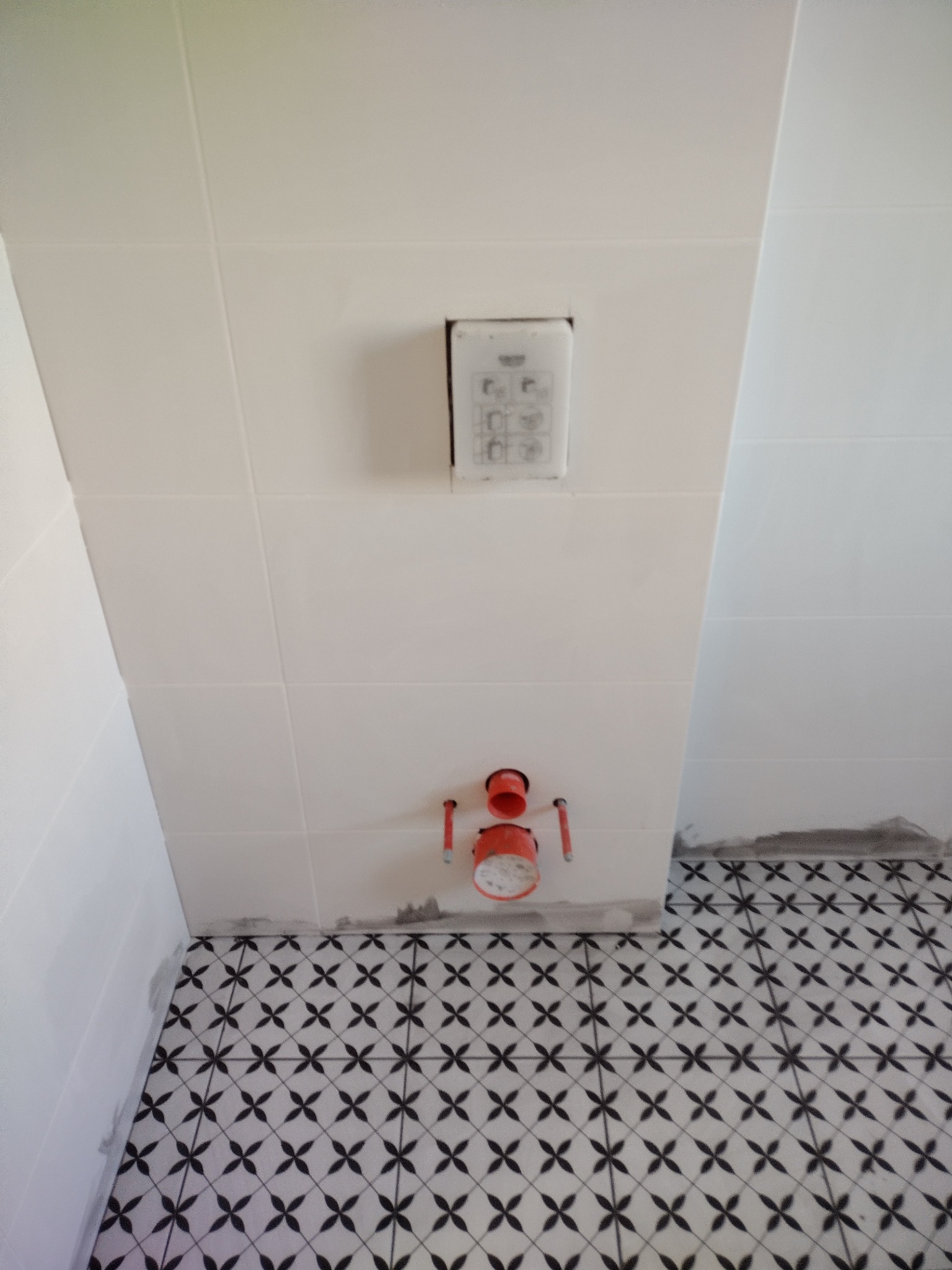 Białe płytki ścienne z zamontowanym panelem i instalacją sanitarną nad czarno-białą podłogą w geometryczny wzór. Widoczne ślady po fugowaniu.