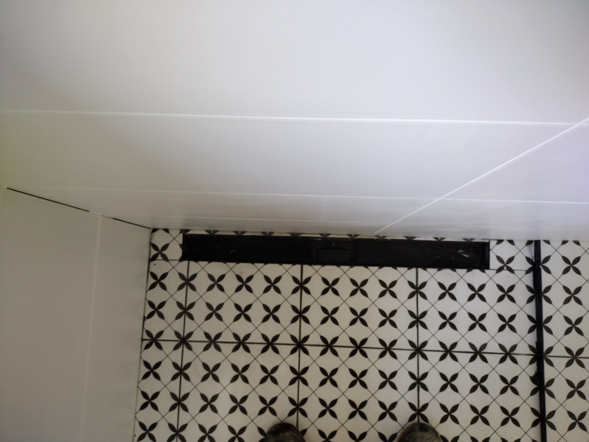 Biała ściana i sufit kontrastują z czarno-białą podłogą w geometryczny wzór. Widoczny czarny odpływ liniowy i fragment butów w dolnej części kadru.