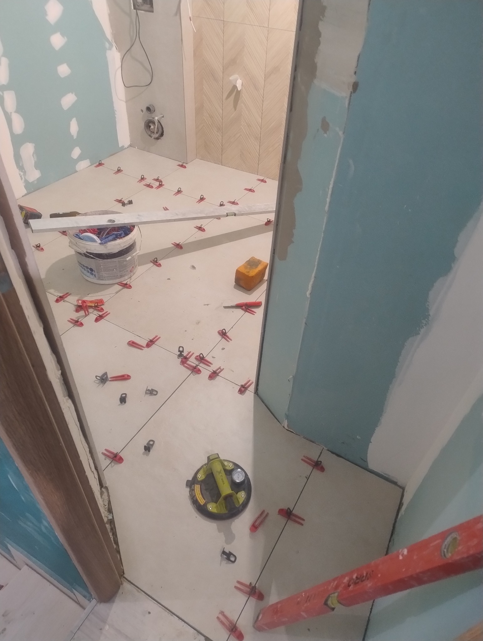 Układanie jasnych płytek podłogowych w łazience, widoczne narzędzia glazurnicze: przyssawka, poziomica, krzyżyki dystansowe. Ściany przygotowane do dalszych prac.