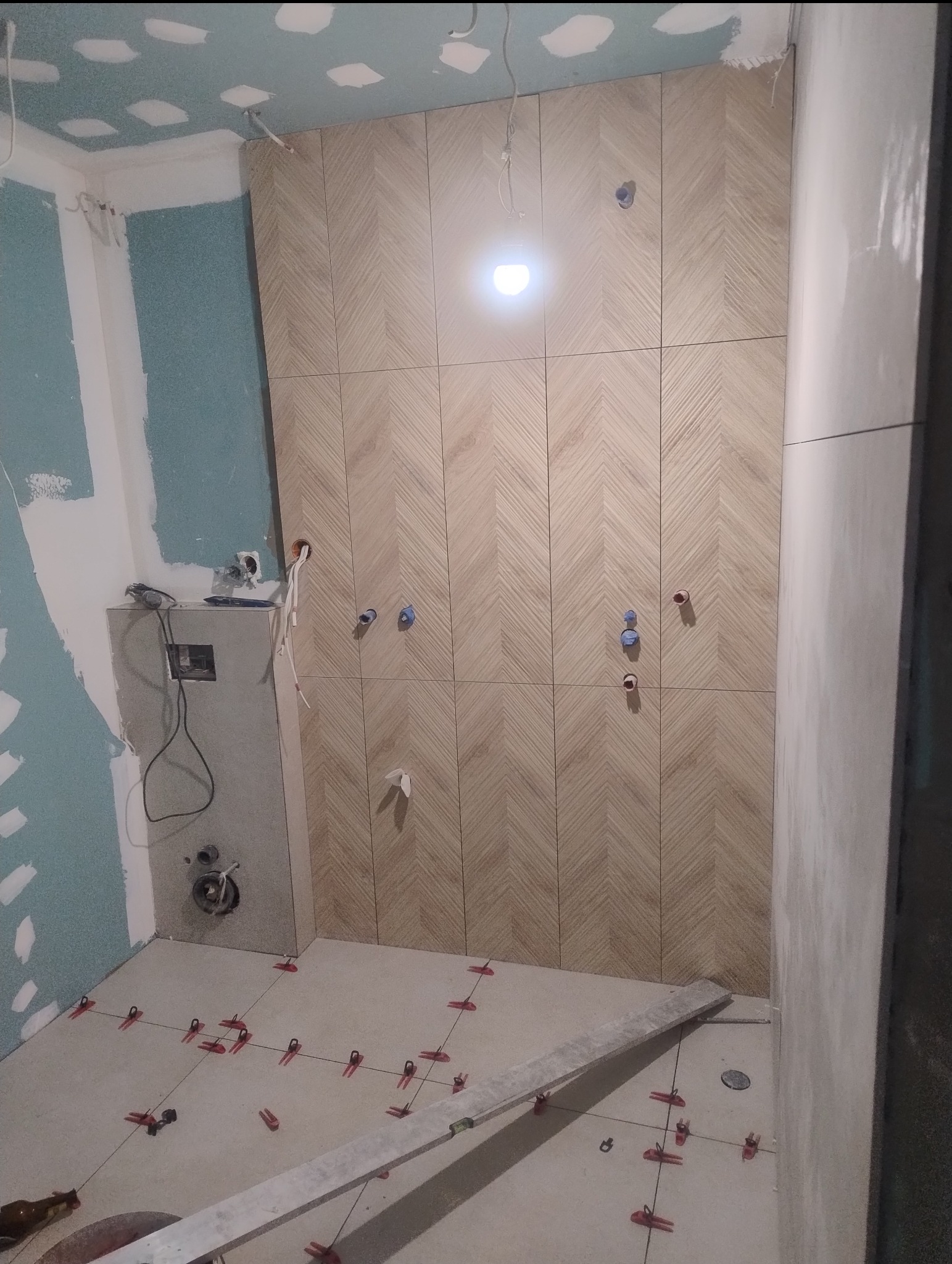 Montaż płytek chevron na ścianie w łazience, widoczne krzyżyki dystansowe na podłodze i niezakończone prace wykończeniowe.