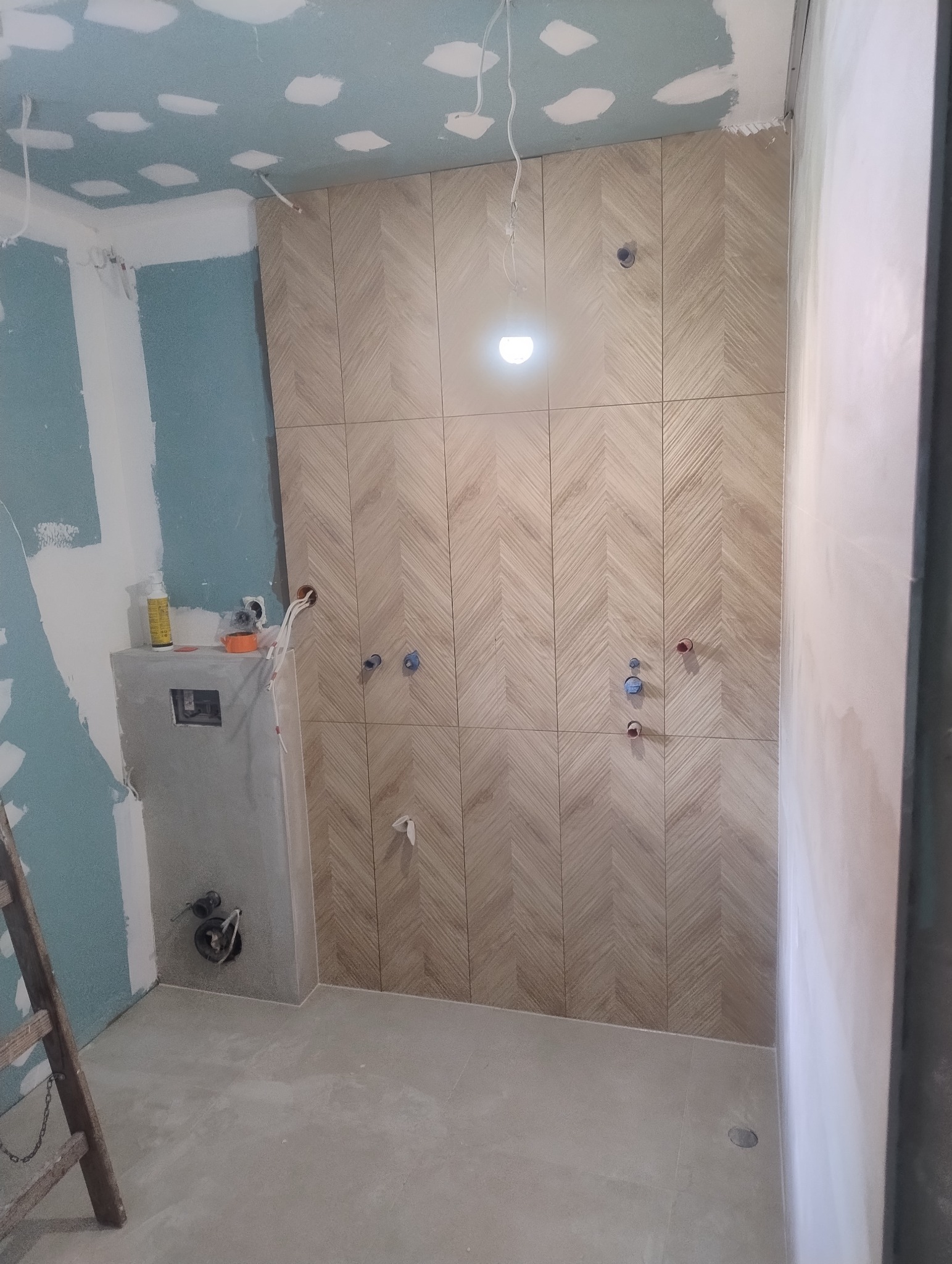 Ułożone płytki w jodełkę na ścianie w łazience w trakcie remontu. Widoczne wystające rury i instalacje. Na suficie płyty gipsowe ze śladami szpachlowania.