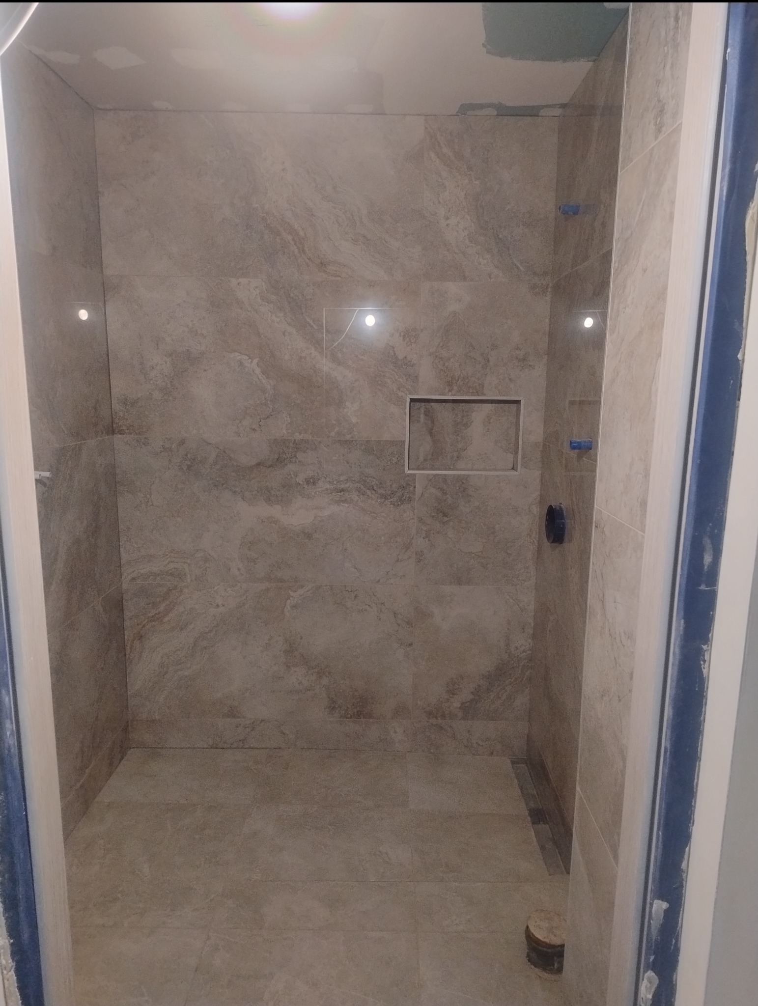 Wykończona kabina prysznicowa z beżowymi płytkami imitującymi marmur, wnęką na kosmetyki i odpływem liniowym. Widoczne elementy montażowe i niedokończone wykończenia.