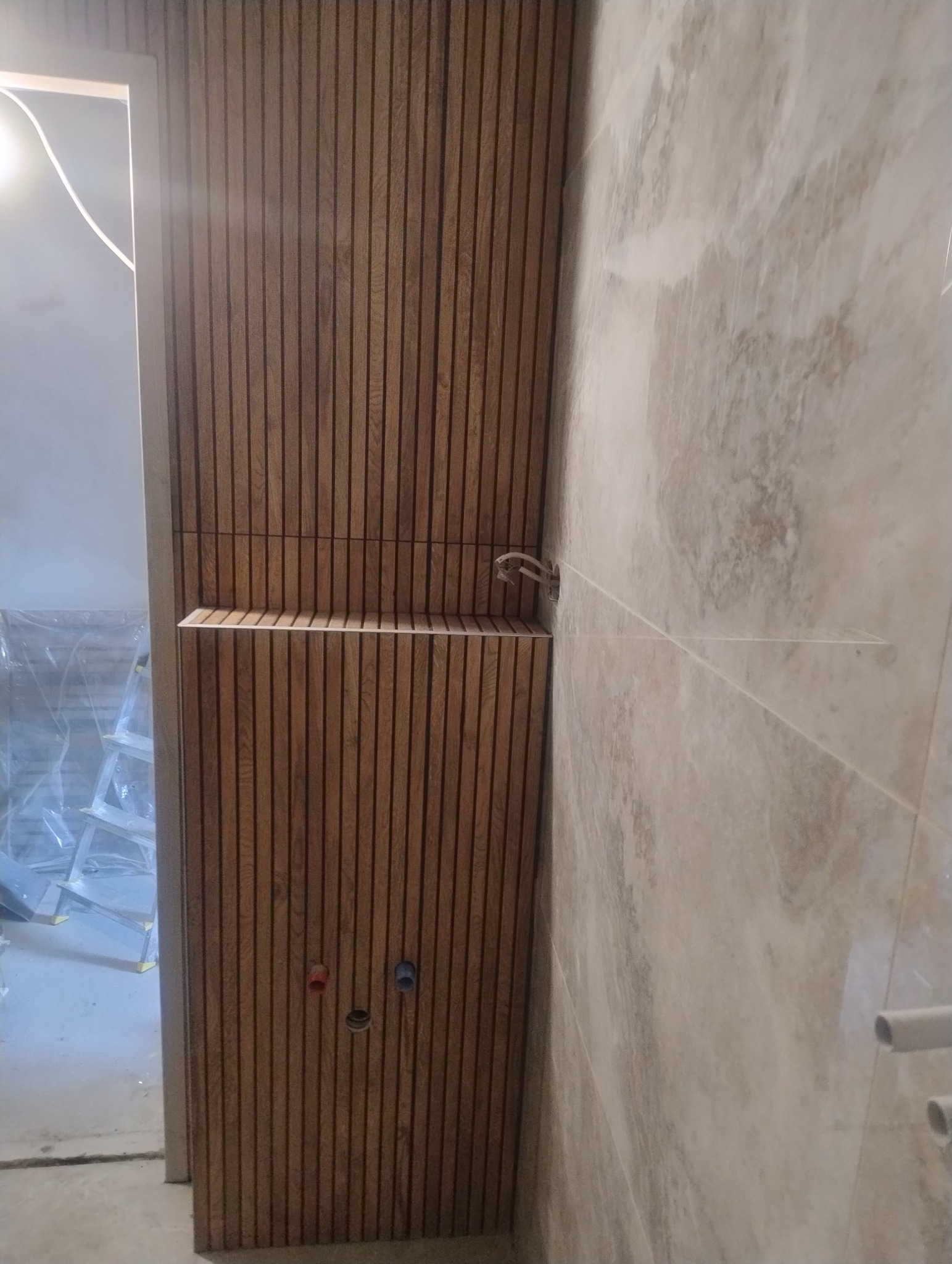 Drewniana boazeria lamelowa w łazience, z widocznymi otworami na instalacje i częściowo położonymi płytkami. W tle widoczna drabina i folia zabezpieczająca.