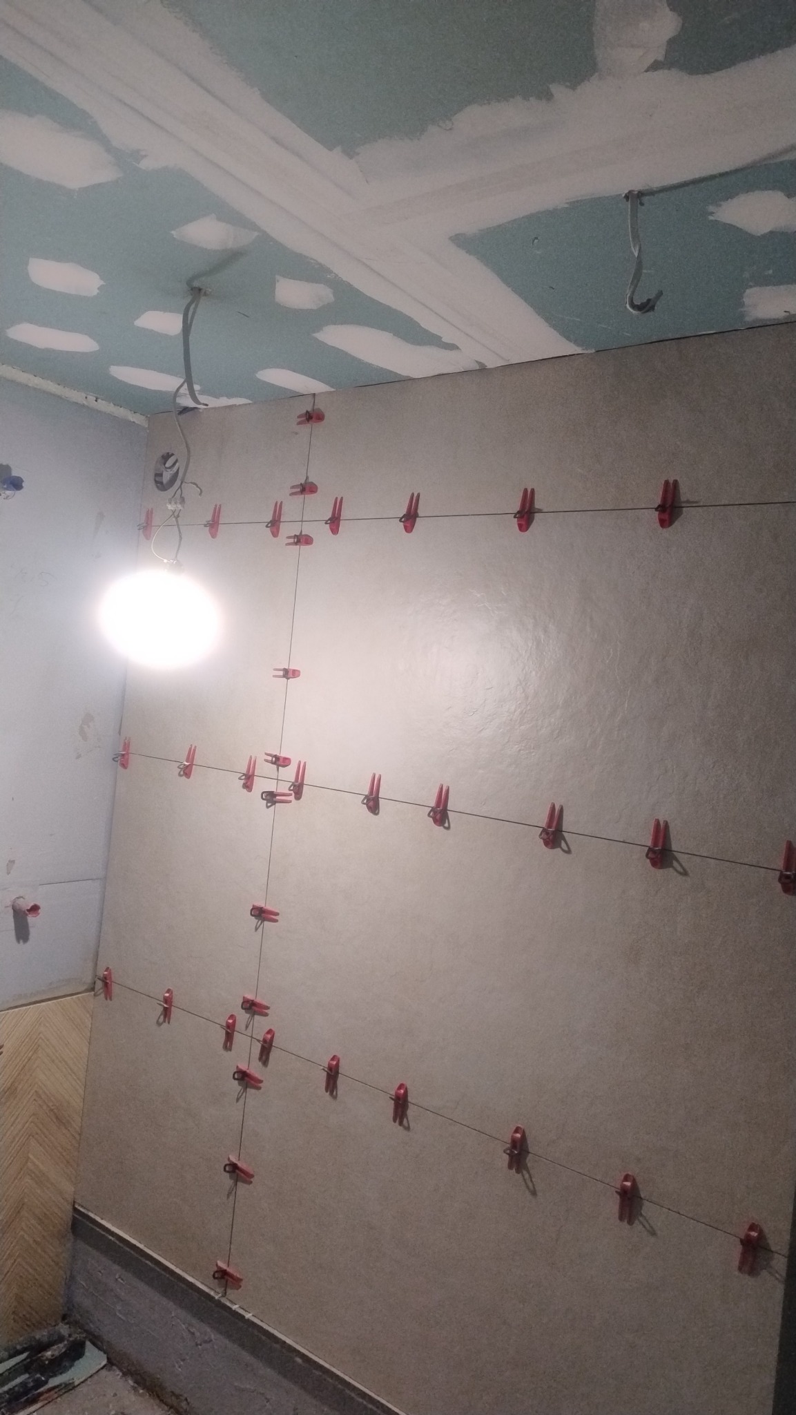 Ściana z jasnymi płytkami, w trakcie montażu, z użyciem klipsów poziomujących i sznurka. Widoczny fragment sufitu z płyt gipsowych i instalacją elektryczną.