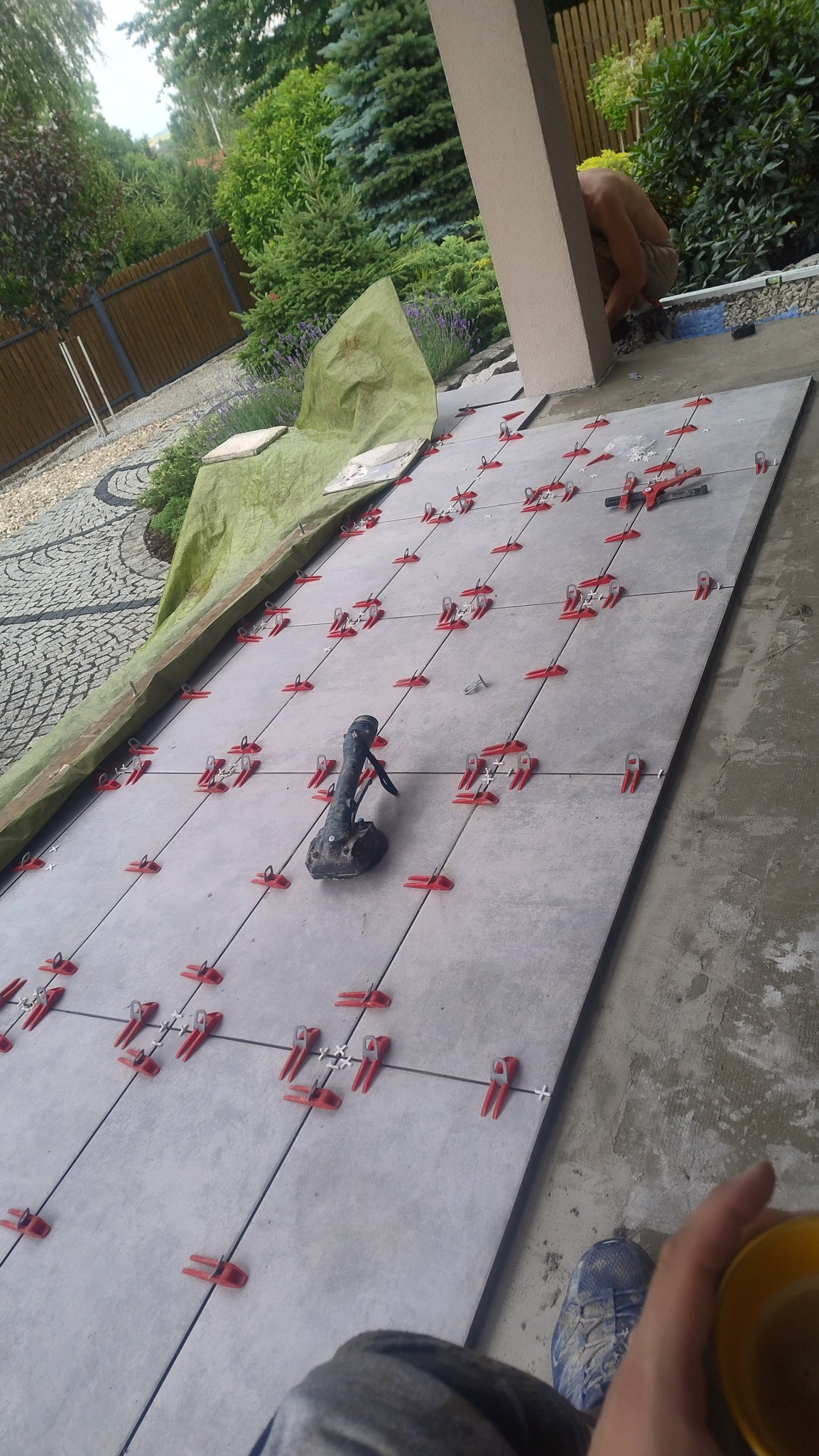 Układanie płytek na zewnątrz z użyciem systemu poziomowania. Widoczna szlifierka kątowa i fragment ogrodu w tle. Praca w toku, widoczne detale mocowania.