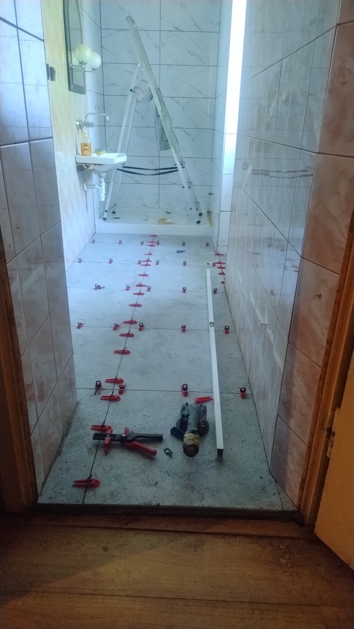 Łazienka w trakcie układania płytek. Widoczne narzędzia, poziomica i krzyżyki dystansowe.  Umywalka i drabina w tle.