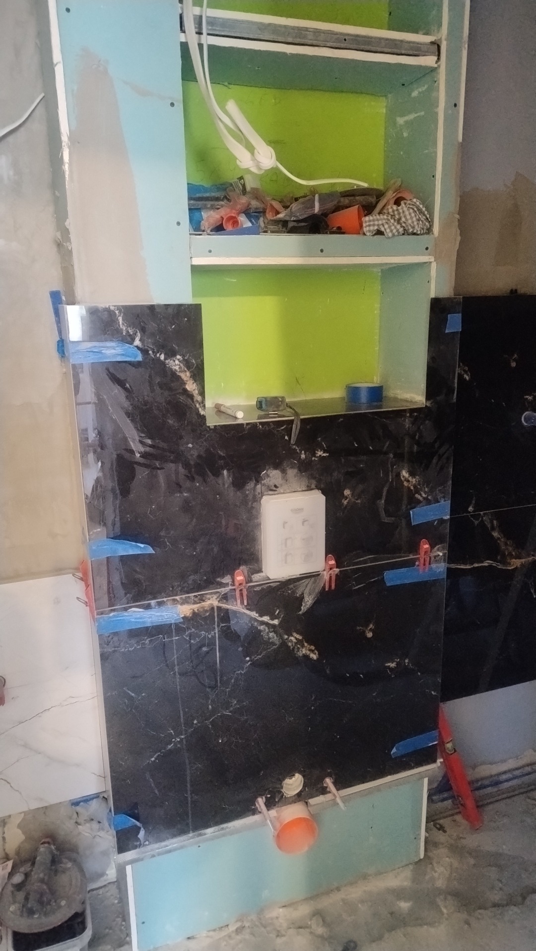 Montaż czarnych, marmurkowych płytek w łazience z wnękami. Widoczne narzędzia i materiały budowlane, niebieska taśma i poziomica. Etap pracy w toku.