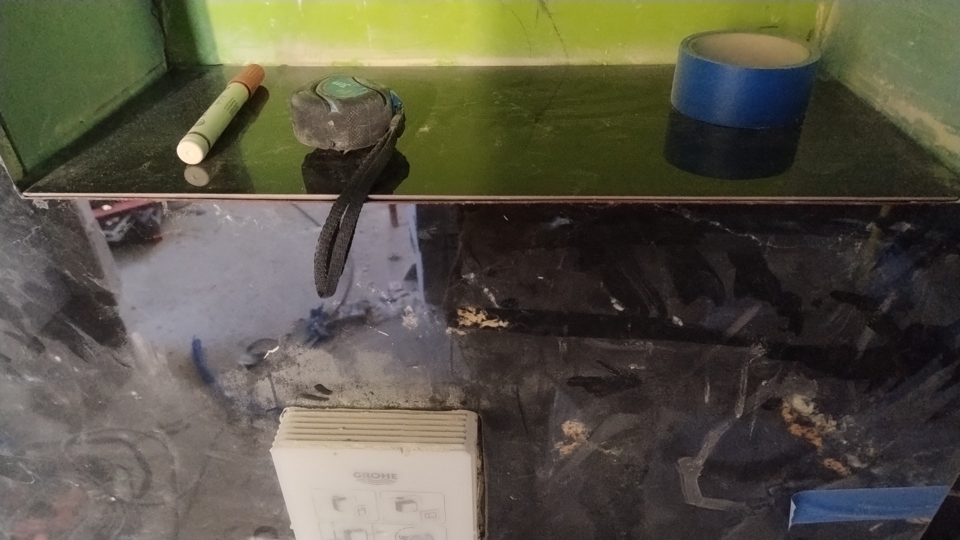 Fragment łazienki w trakcie prac wykończeniowych: czarna, błyszcząca glazura, narzędzia (miarka, marker), taśma malarska i przycisk spłuczki Grohe.