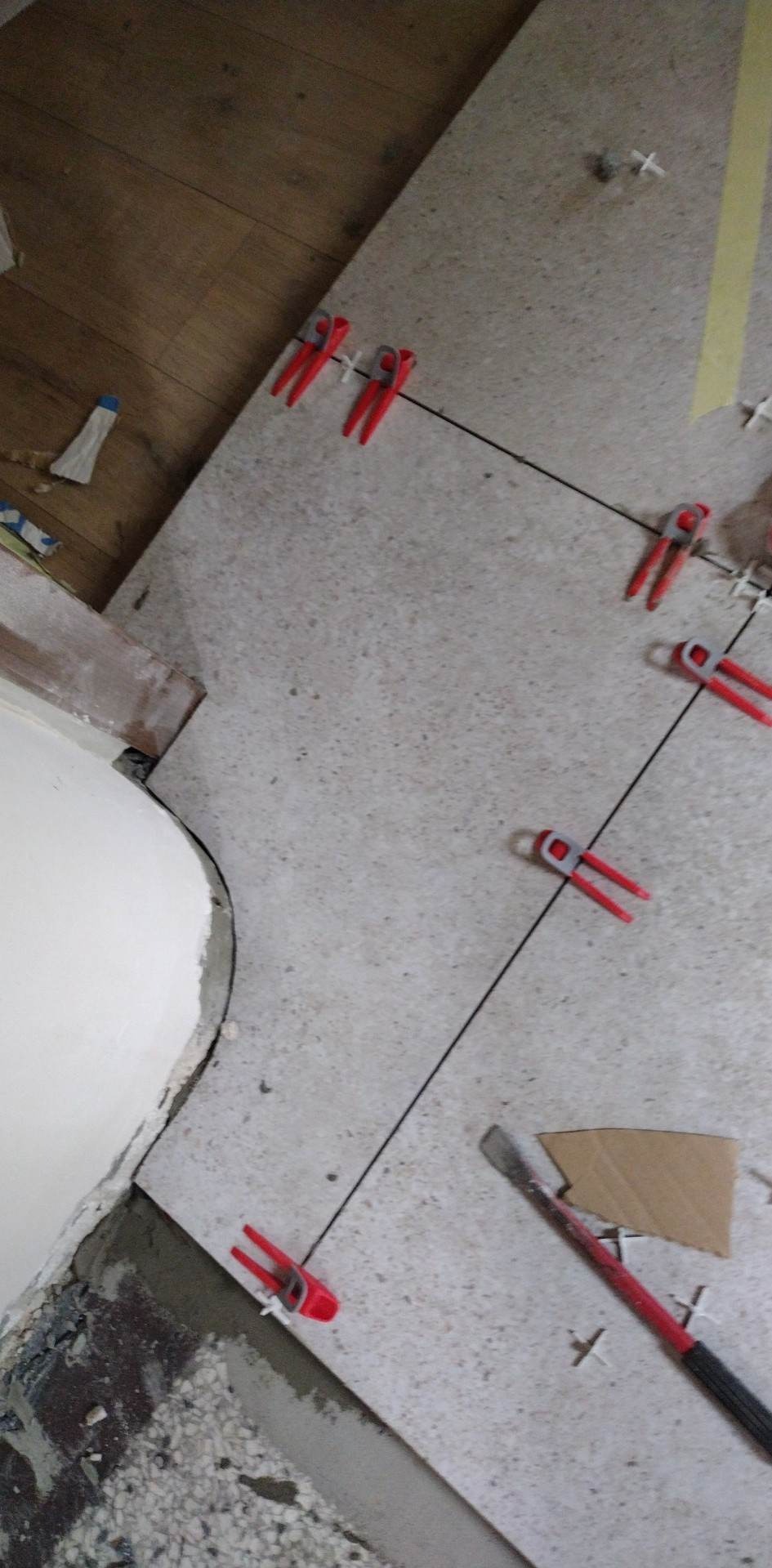 Układanie jasnych płytek podłogowych z użyciem systemu poziomowania płytek (czerwone klipsy i czarne kliny). Widoczne krzyżyki dystansowe i fragment drewnianej podłogi.