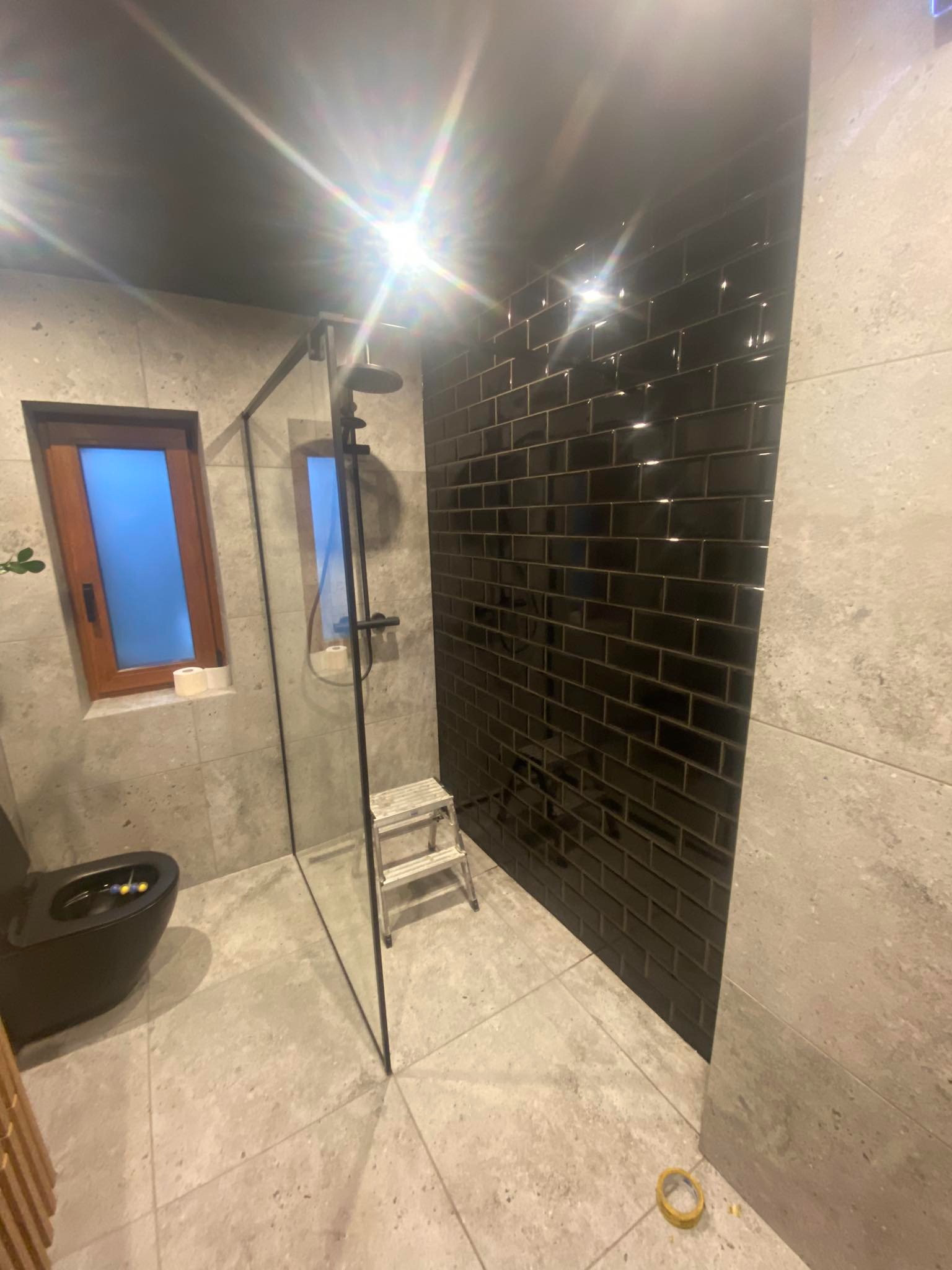 Nowoczesna łazienka z czarną armaturą i błyszczącymi, czarnymi płytkami w stylu cegiełek na ścianie kabiny prysznicowej. Widoczny fragment toalety i okno.