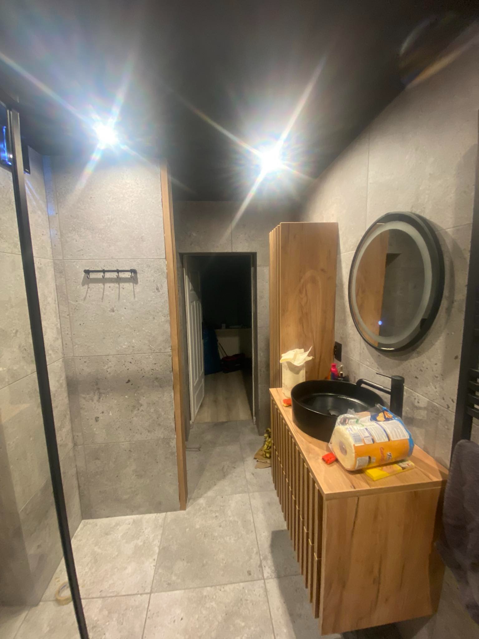 Nowoczesna łazienka z szarą płytką, drewnianą szafką pod umywalką i okrągłym lustrem z podświetleniem. Widoczny fragment kabiny prysznicowej.