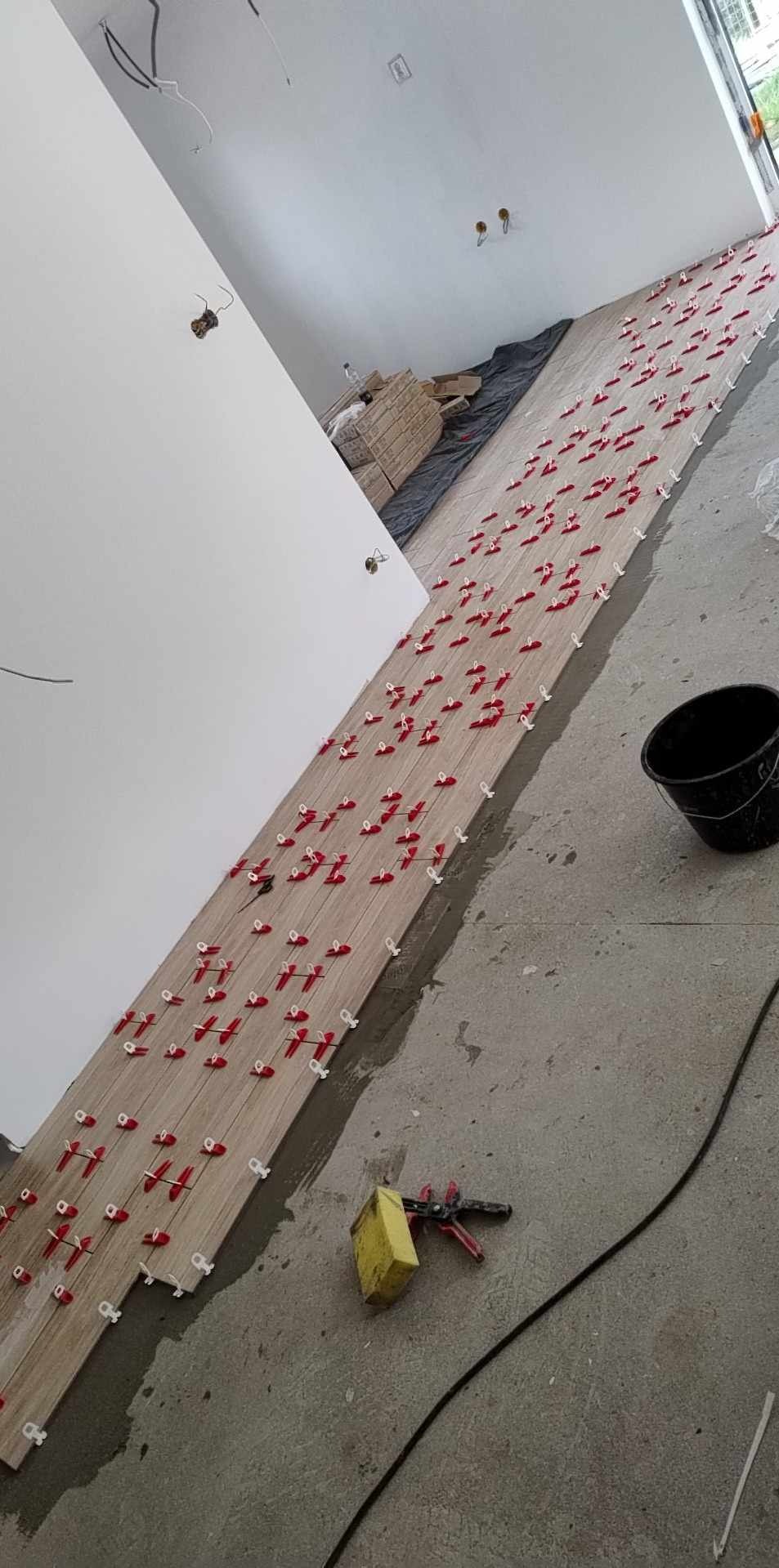 Układanie płytek podłogowych z użyciem klipsów poziomujących. Widoczny fragment ściany, narzędzia glazurnicze i wiadro. Praca w toku.