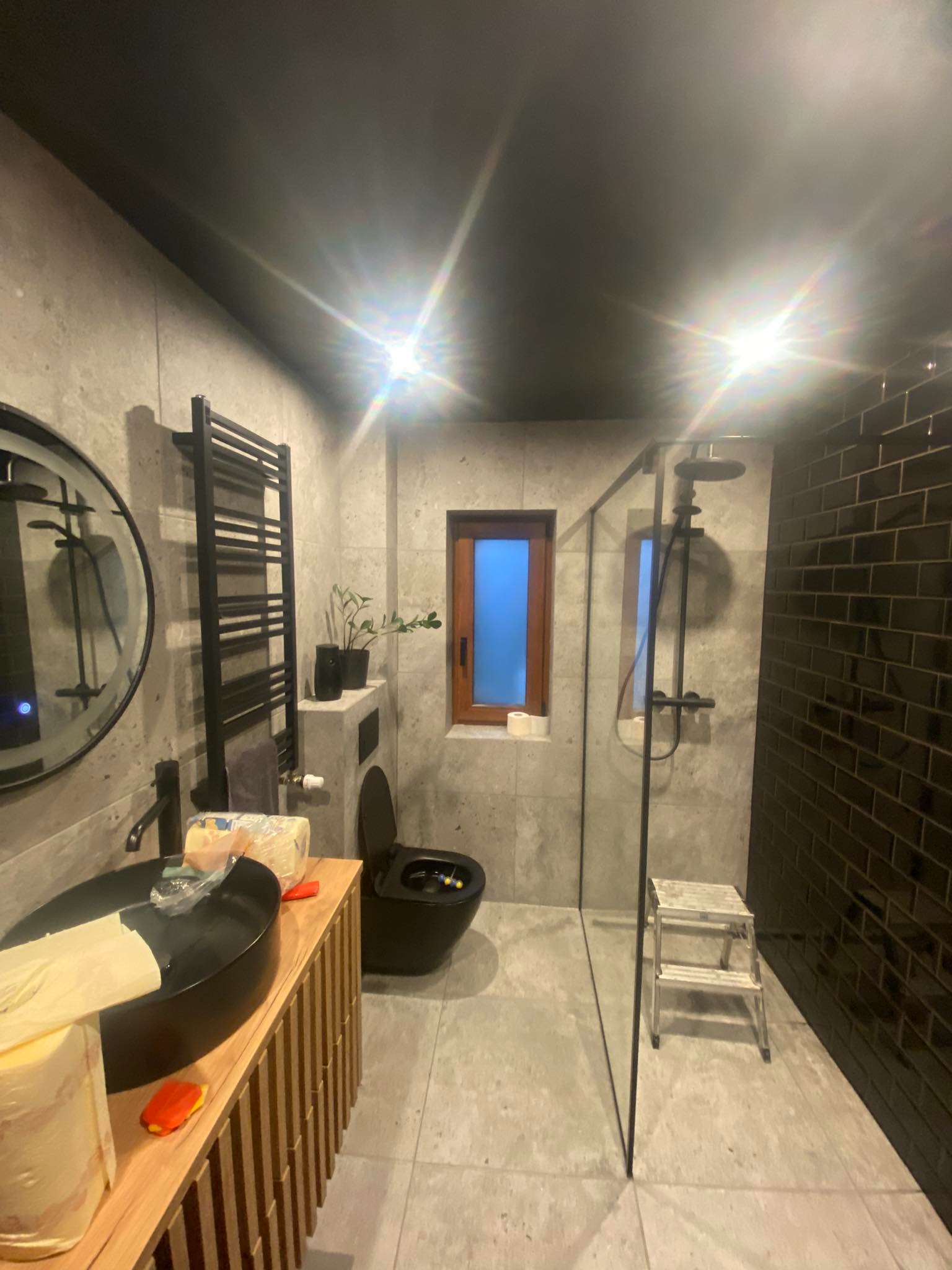 Nowoczesna łazienka z czarną armaturą: umywalka, grzejnik drabinkowy, toaleta. Ściany wyłożone szarą płytką, kabina prysznicowa ze szklaną ścianą i czarną glazurą.