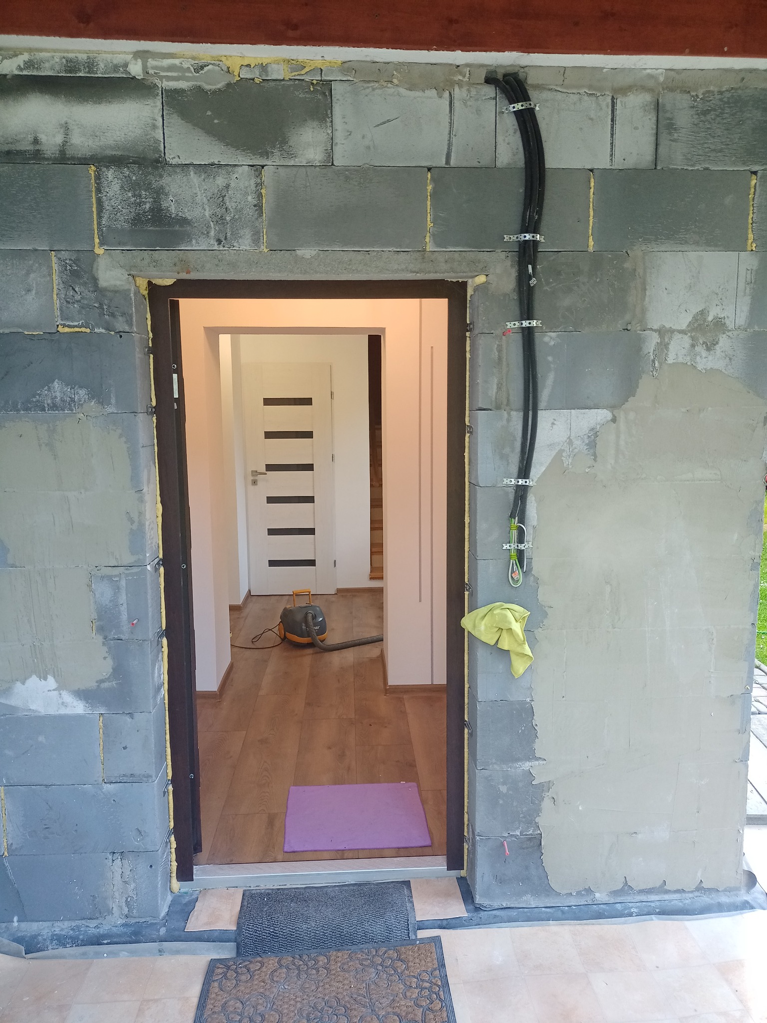 Wnętrze w trakcie wykańczania: surowe ściany z betonu komórkowego, widoczne przewody elektryczne, zamontowane ościeżnice i częściowo wykończona podłoga z odkurzaczem.