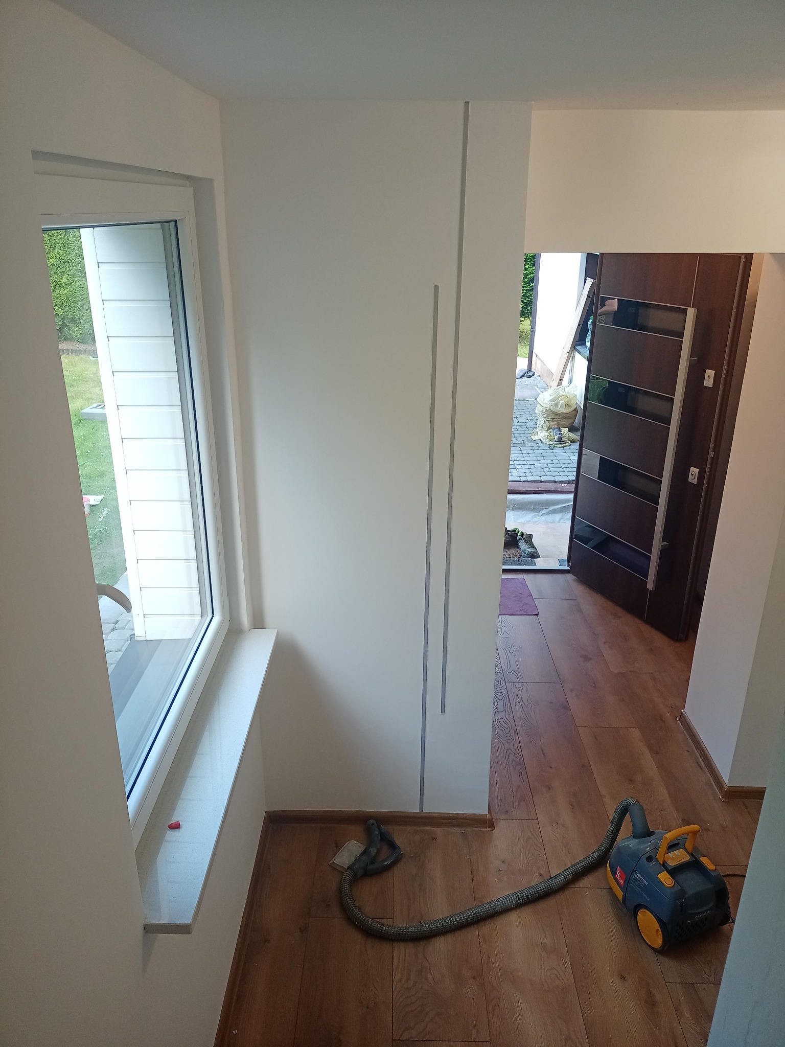 Jasne wnętrze po remoncie: białe ściany z subtelnymi szarymi akcentami, drewnopodobna podłoga i okno z widokiem na zieleń. Odkurzacz na podłodze sugeruje prace porządkowe.