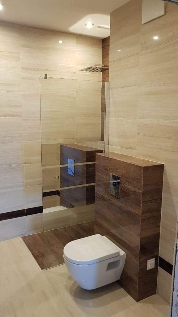 Nowoczesna łazienka z beżowymi płytkami imitującymi marmur, kabiną prysznicową typu walk-in z przezroczystą szybą, podwieszaną toaletą i obudową stelaża WC w okleinie drewnopodobnej.