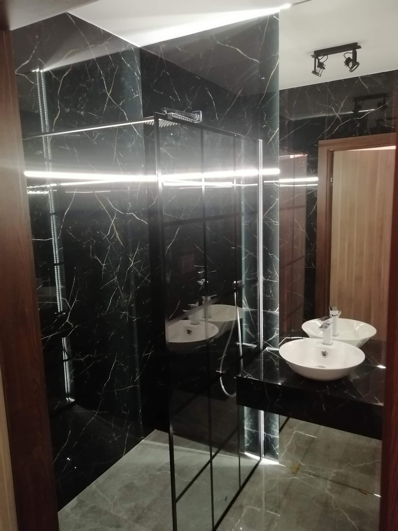 Nowoczesna łazienka z czarnymi marmurowymi płytkami, podświetleniem LED i szklaną kabiną prysznicową typu walk-in, odbicie dwóch białych umywalek nablatowych w lustrze.