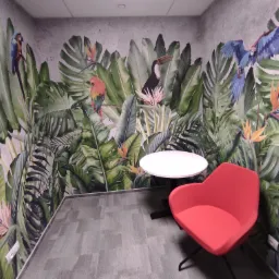Pokój z tropikalną fototapetą przedstawiającą liście, papugi i tukana, z okrągłym białym stolikiem i czerwonym krzesłem.