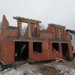 Wznoszenie ścian zewnętrznych z czerwonej cegły porotherm na budowie domu jednorodzinnego w zimowej scenerii, widoczna izolacja fundamentów i częściowo wymurowane nadproża.