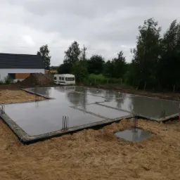 Wylany fundament pod dom jednorodzinny z wystającymi prętami zbrojeniowymi, widoczny samochód betoniarka oraz przyczepa kempingowa w tle.