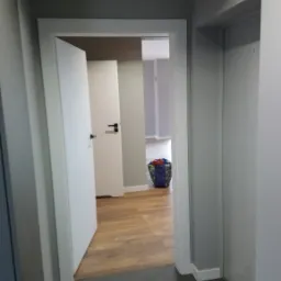 Szare wnętrze mieszkania po remoncie, widok na drewnianą podłogę przez otwarte białe drzwi, na podłodze torba z narzędziami.