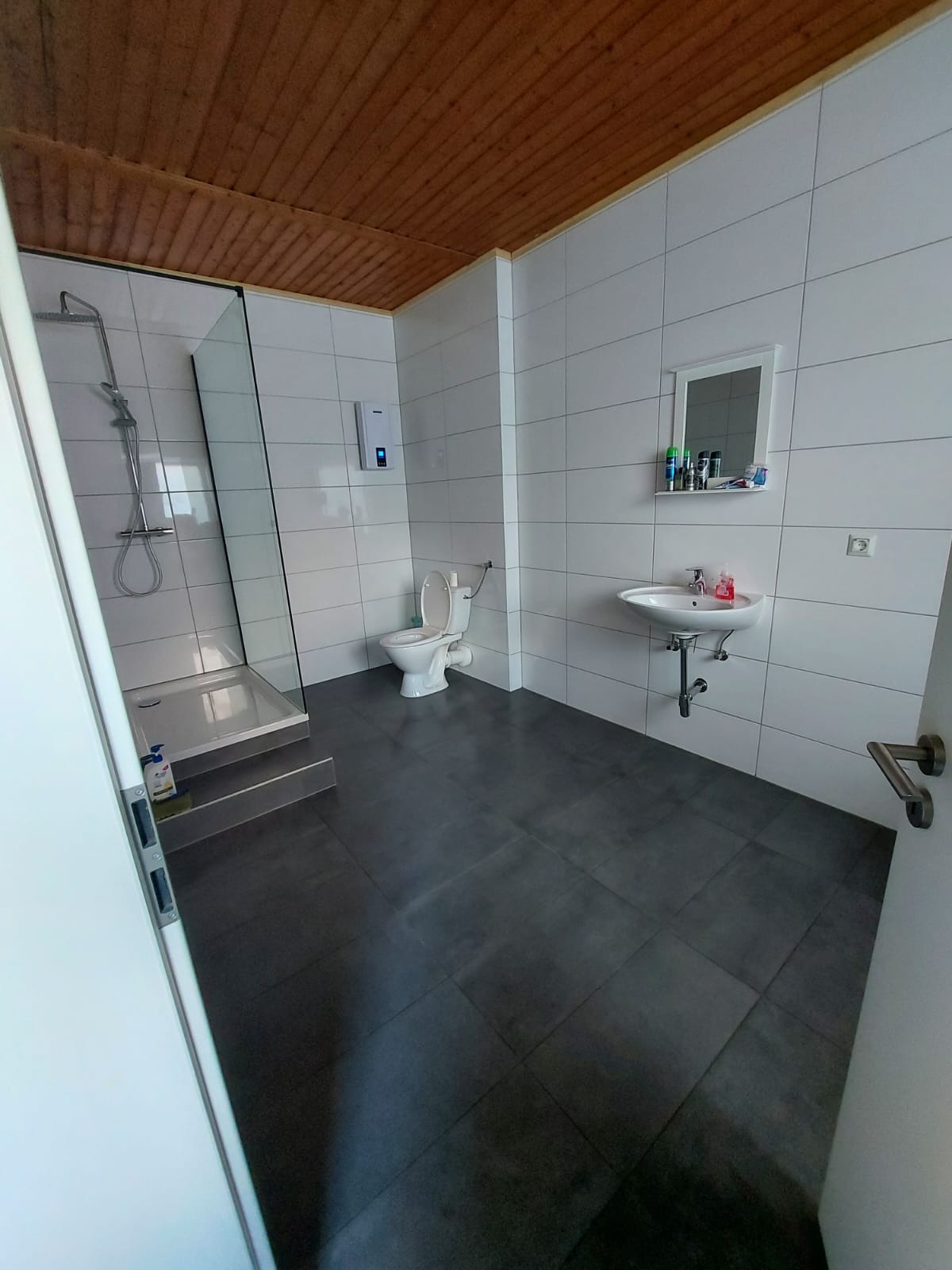 Wnętrze łazienki po remoncie: kabina prysznicowa ze szklaną ścianką, toaleta, umywalka z szafką, białe płytki na ścianach i ciemnoszara terakota na podłodze, drewniana boazeria na suficie.