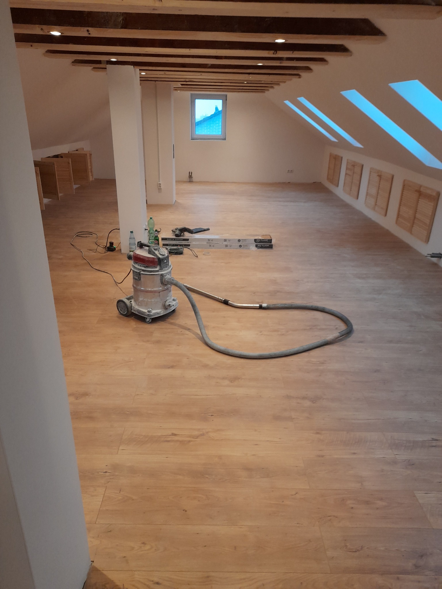 Poddasze po remoncie z nową drewnianą podłogą, widoczny odkurzacz przemysłowy i resztki materiałów budowlanych.