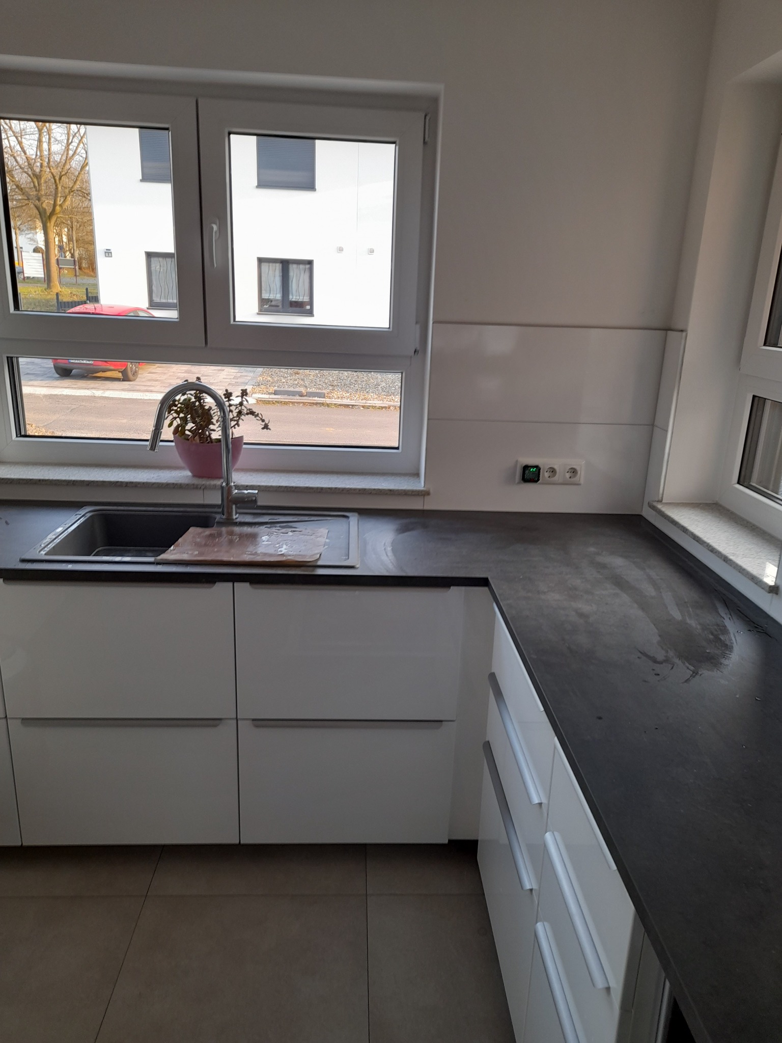 Nowoczesna, minimalistyczna kuchnia z białymi szafkami i ciemnym blatem, z widokiem na budynek mieszkalny za oknem. Na blacie przy zlewie stoi doniczka z rośliną.