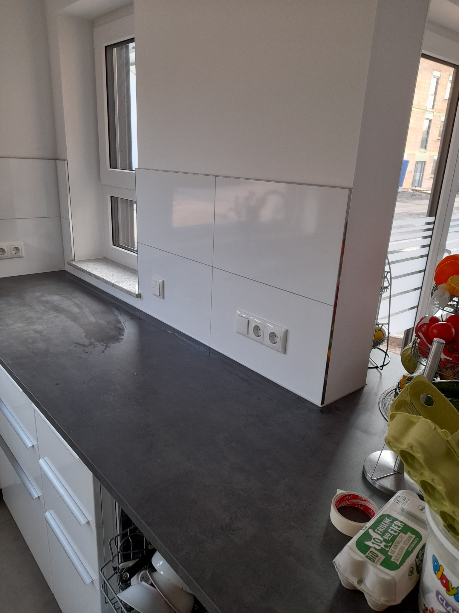 Fragment nowoczesnej kuchni z szarym blatem, białymi szafkami, oknem i białymi panelami ściennymi z gniazdkami elektrycznymi, widoczne akcesoria kuchenne i produkty spożywcze.