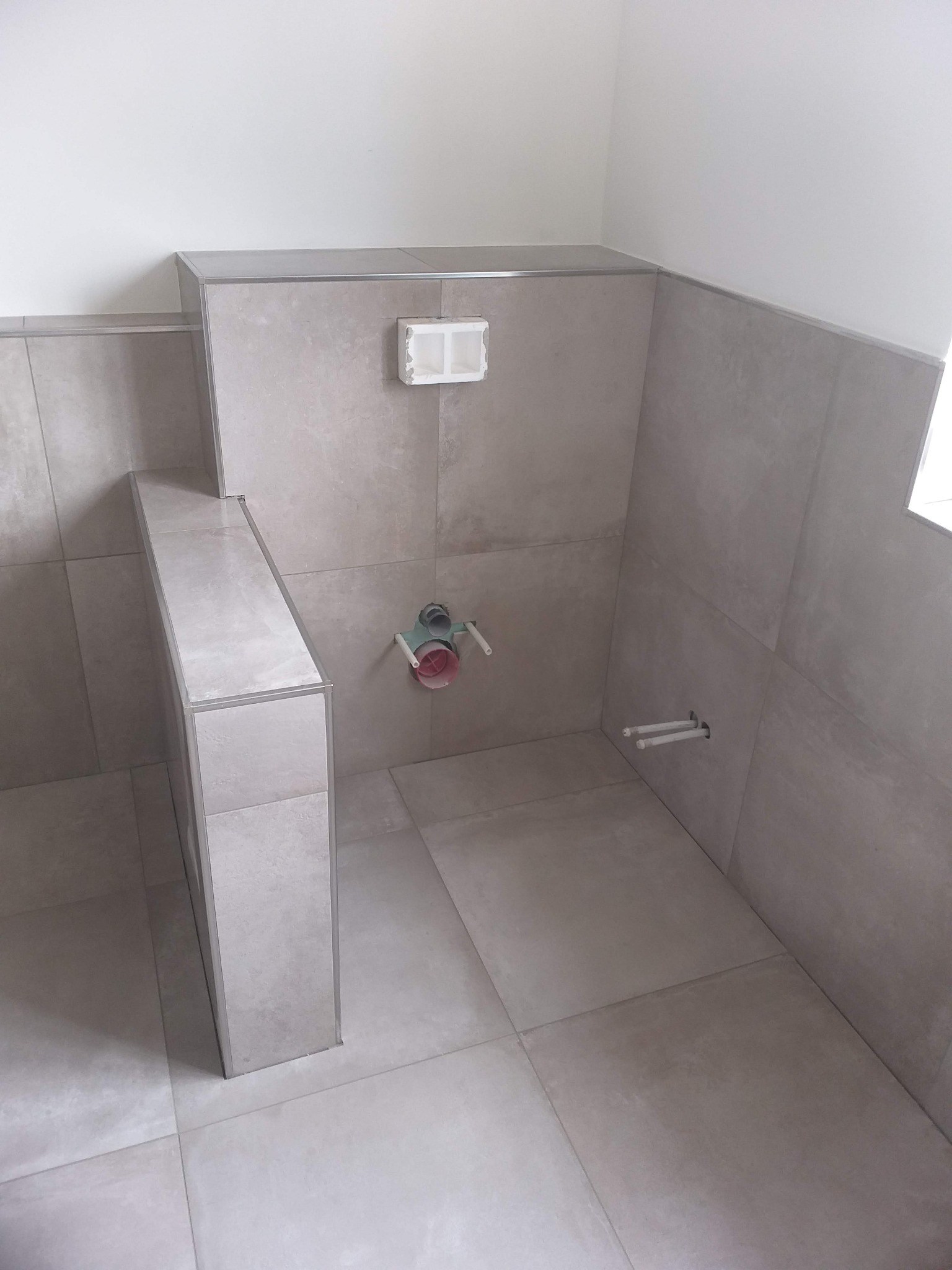 Wykończona łazienka w szarych płytkach z widocznymi przyłączami wodno-kanalizacyjnymi i stelażem podtynkowym WC.