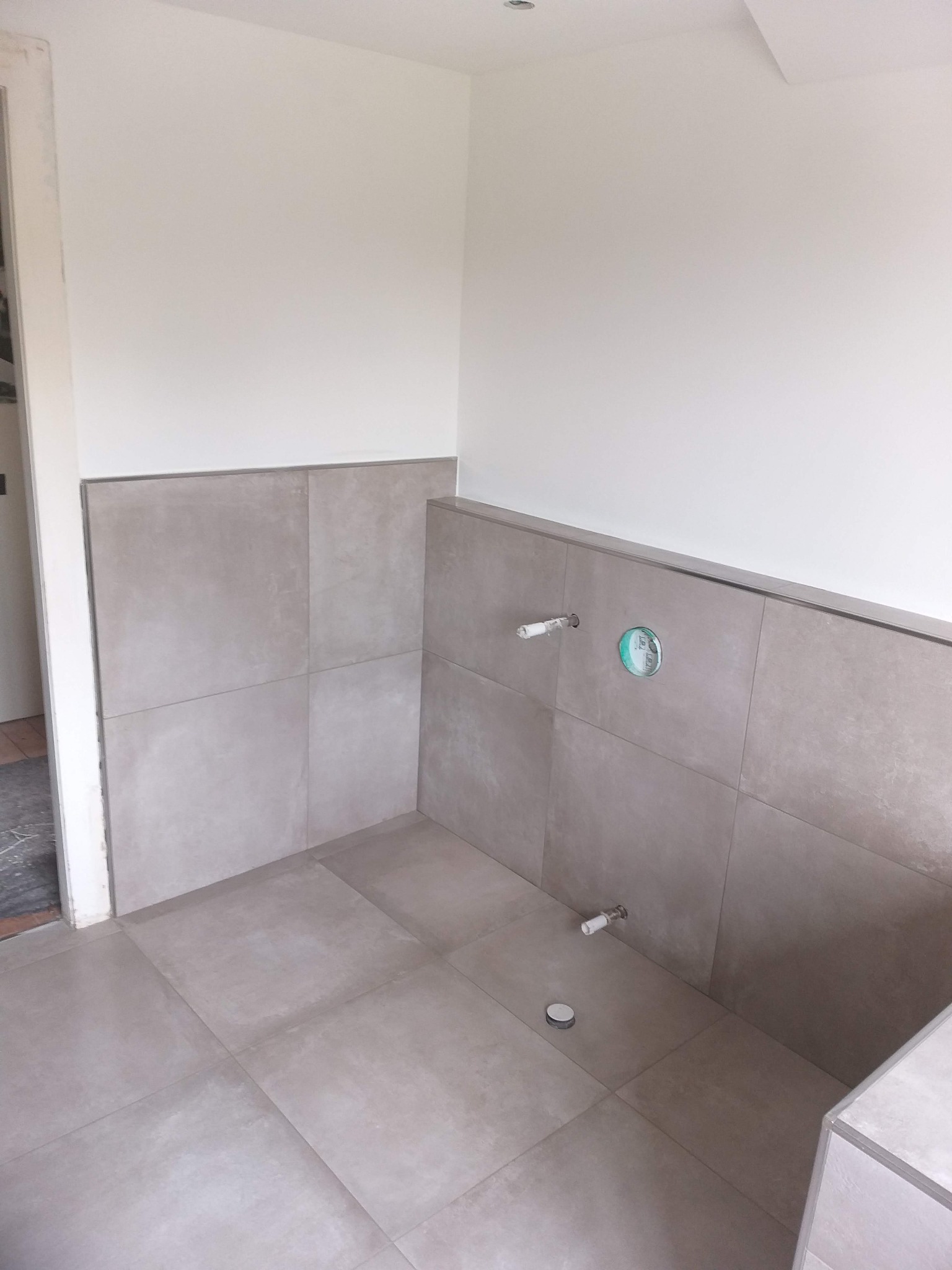 Wykończona dolna część ściany i podłoga w łazience z szarych, kwadratowych płytek ceramicznych, widoczne podejścia wodno-kanalizacyjne.