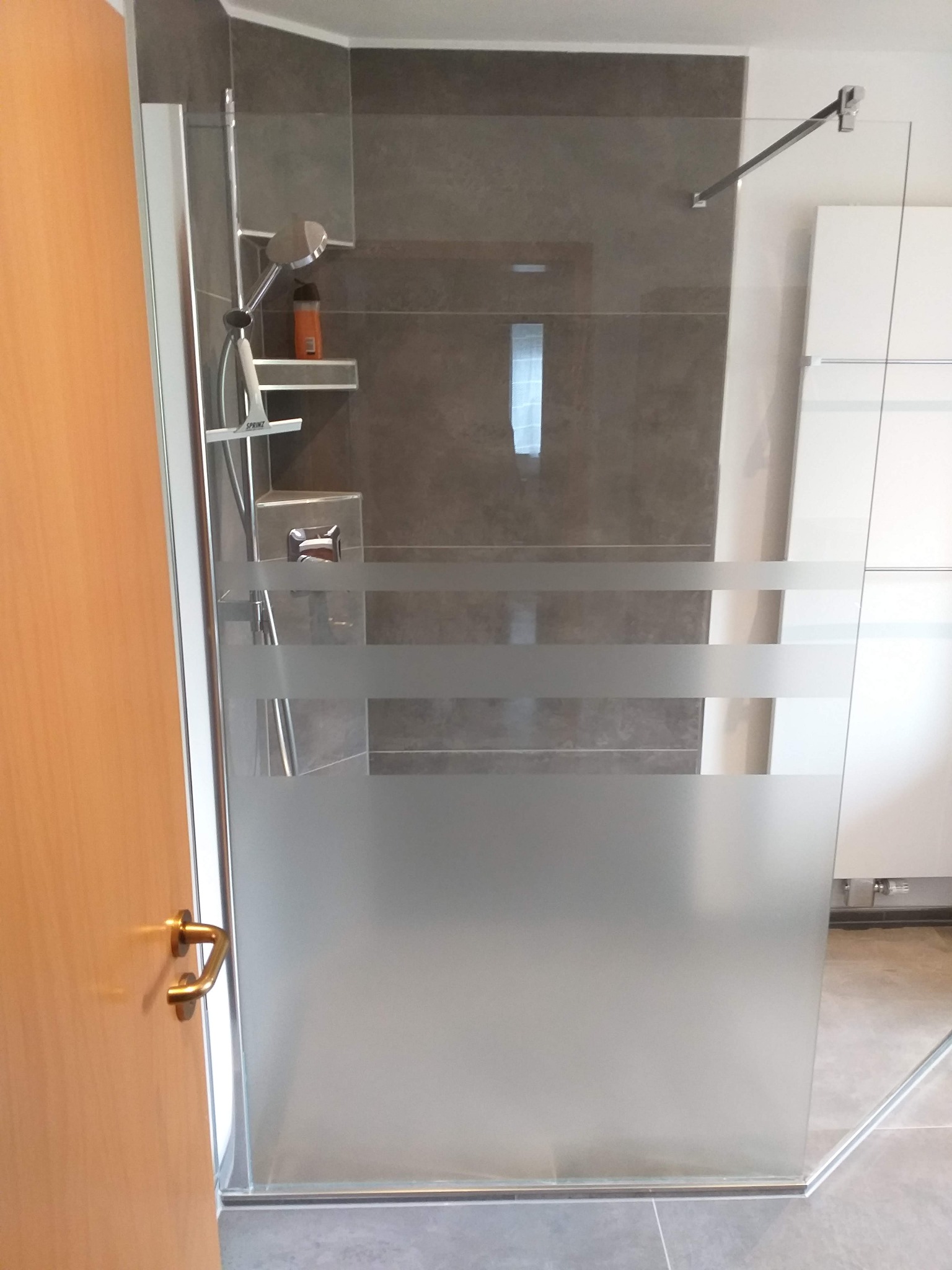 Szklana kabina prysznicowa z matowymi pasami, widoczny panel z szarymi płytkami, bateria prysznicowa, półka z kosmetykami oraz fragment drzwi z klamką.