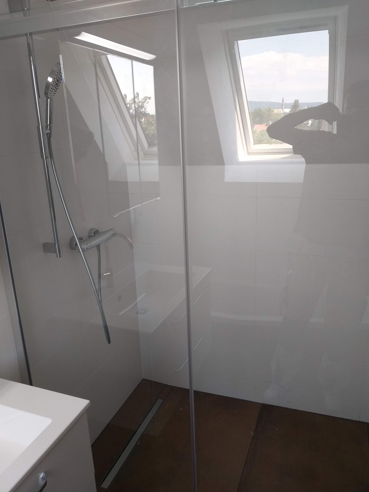 Nowoczesna kabina prysznicowa ze szklaną ścianą, widok na okno dachowe i krajobraz za nim, brązowa podłoga w kabinie, minimalistyczna armatura.