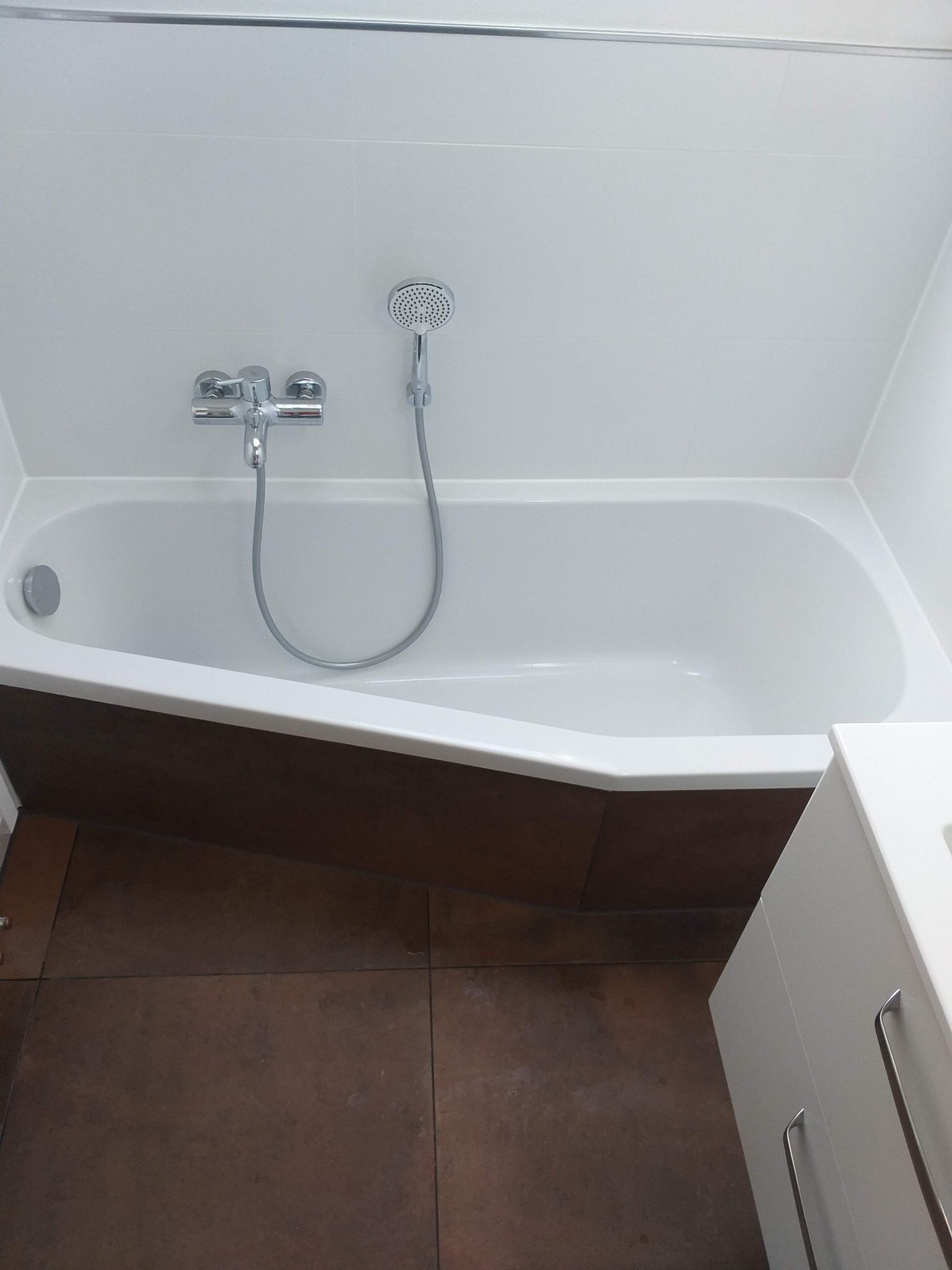 Biała wanna z chromowaną baterią i słuchawką prysznicową na tle białych płytek, z brązową obudową wanny i fragmentem białej szafki łazienkowej, widok z góry.