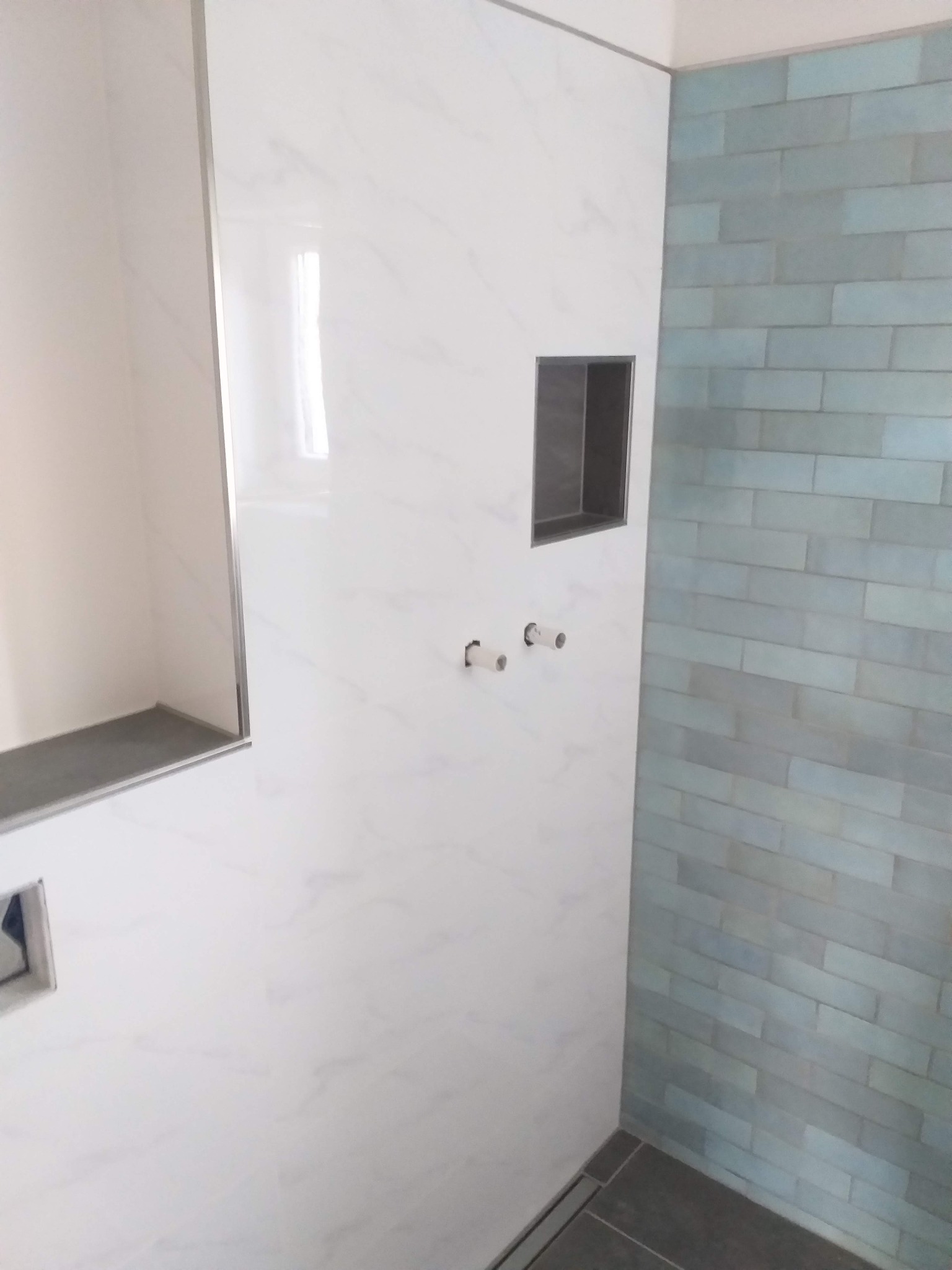 Wnęki w ścianie wyłożonej płytkami imitującymi marmur, obok ściana z niebieskich, podłużnych płytek, widoczne wystające końcówki rur instalacyjnych, szara podłoga.