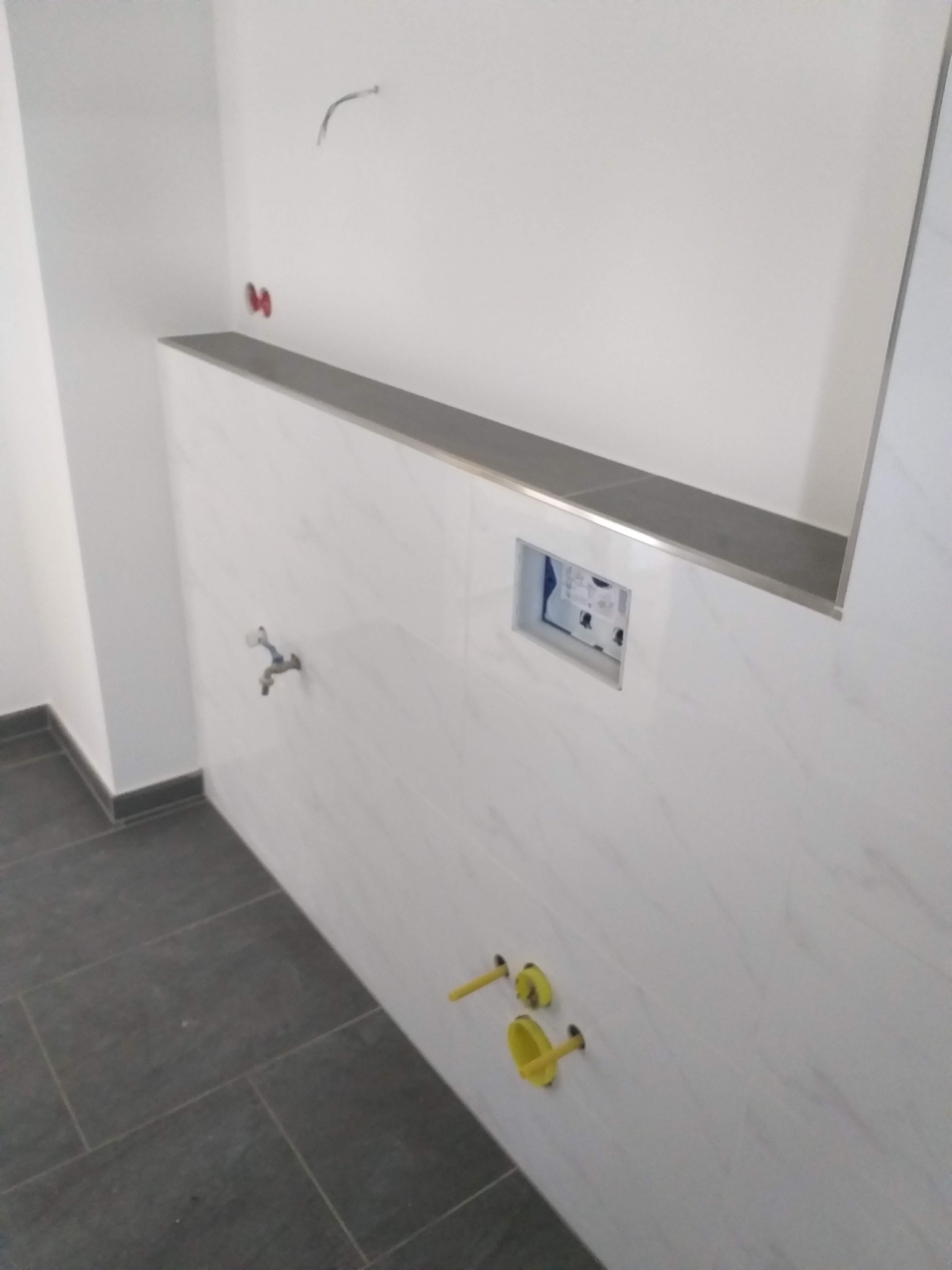 Niedokończona łazienka z białymi marmurkowymi płytkami ściennymi, ciemnoszarą półką wbudowaną w ścianę, przygotowanymi przyłączami wodnymi i szarą podłogą z płytek.