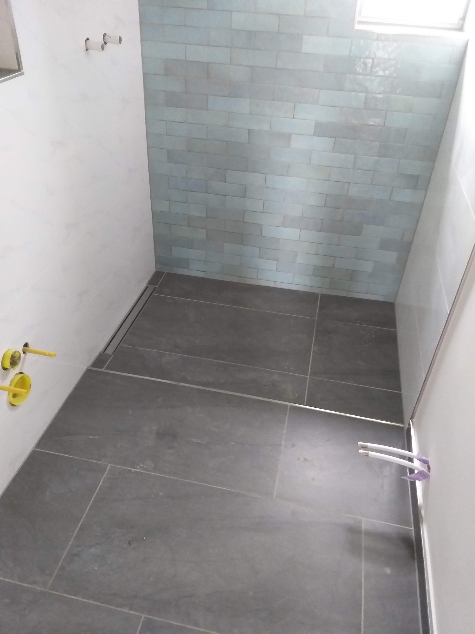 Nowoczesna łazienka w trakcie wykańczania: szare płytki podłogowe, błękitne płytki ścienne ułożone w cegiełkę, widoczne instalacje hydrauliczne i odpływ liniowy.