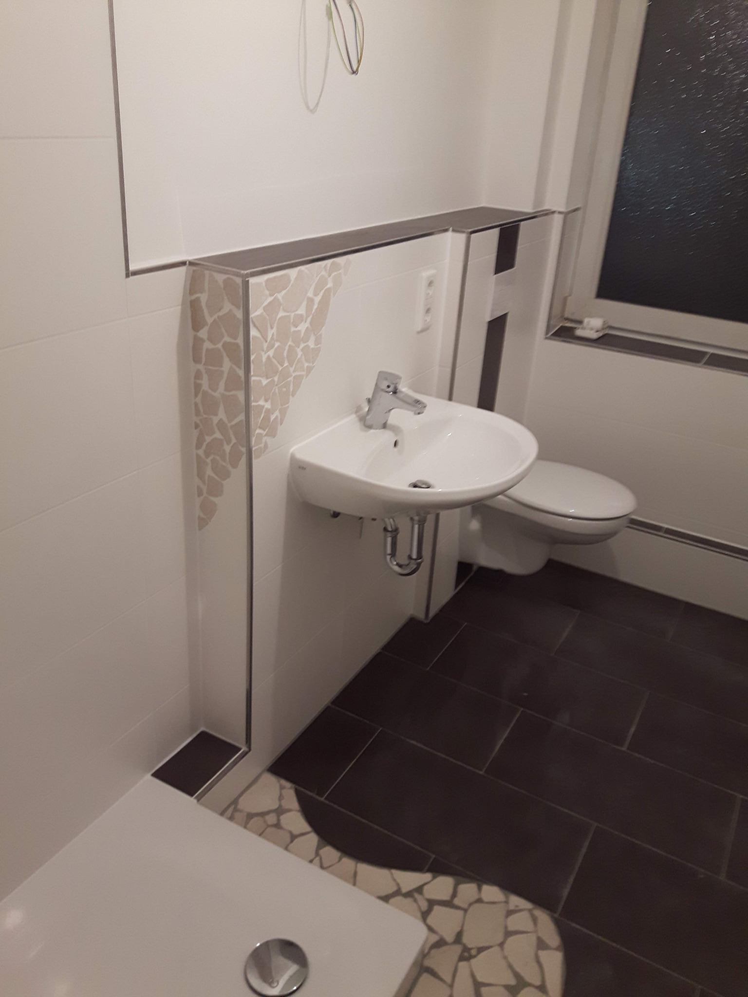 Biała umywalka zamontowana na ścianie z dekoracyjną mozaiką w łazience z ciemnobrązową podłogą i białymi płytkami ściennymi, widoczne fragmenty toalety i kabiny prysznicowej.