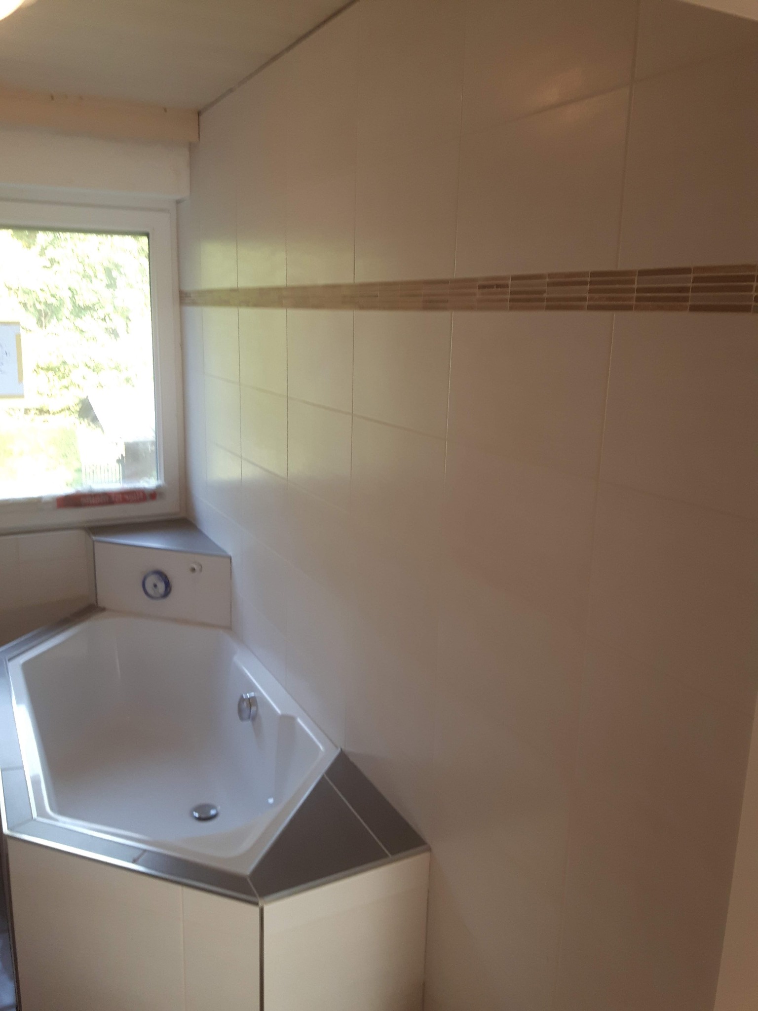 Narożna wanna w wykończonej kafelkami łazience w Kassel, widoczne okno i dekoracyjny pas płytek na ścianie.