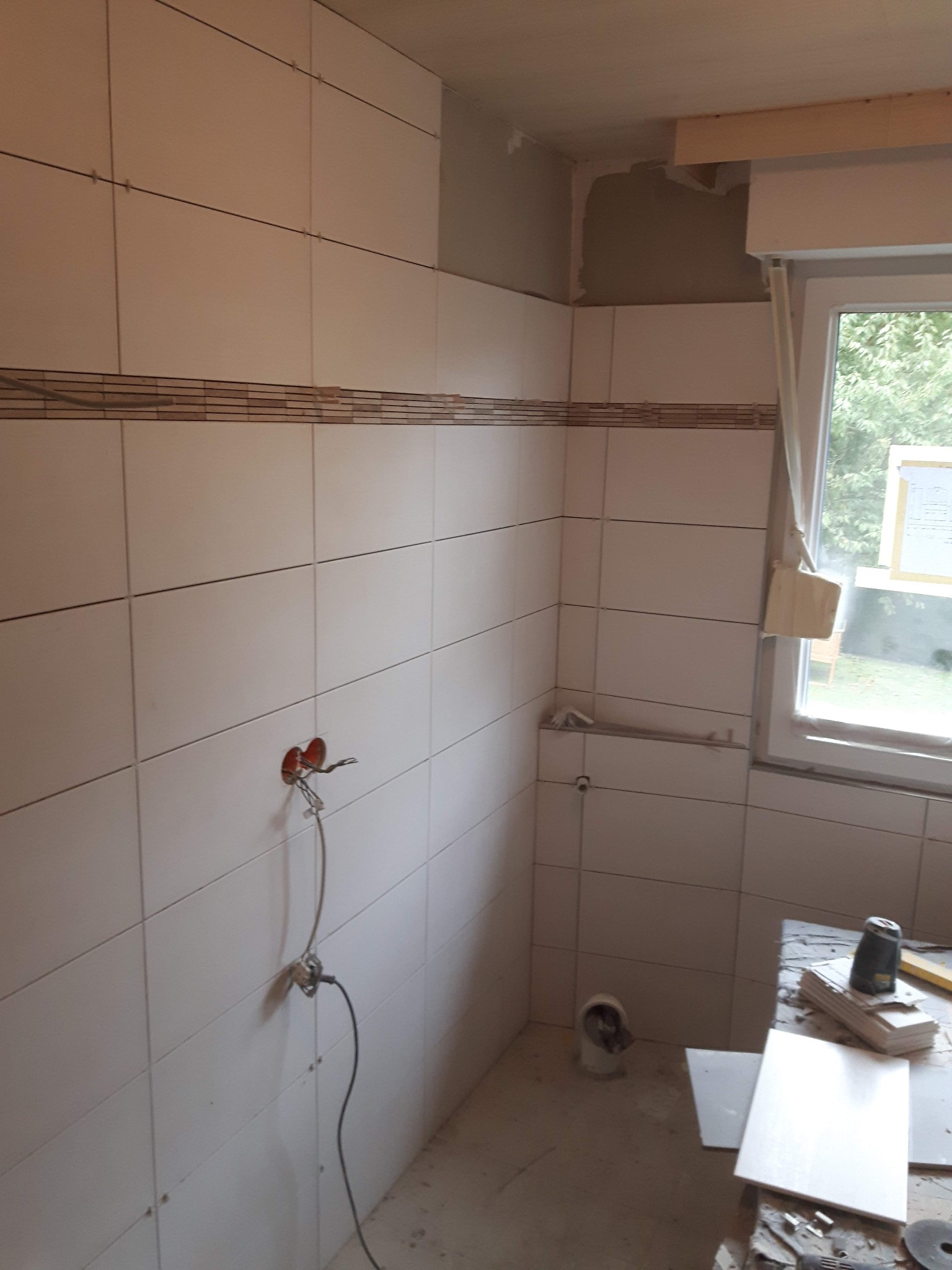 Białe płytki ceramiczne z dekoracyjnym pasem mozaiki ułożone na ścianach łazienki, widoczne wystające kable elektryczne, okno z widokiem na zieleń oraz narzędzia i materiały budowlane na podłodze...
