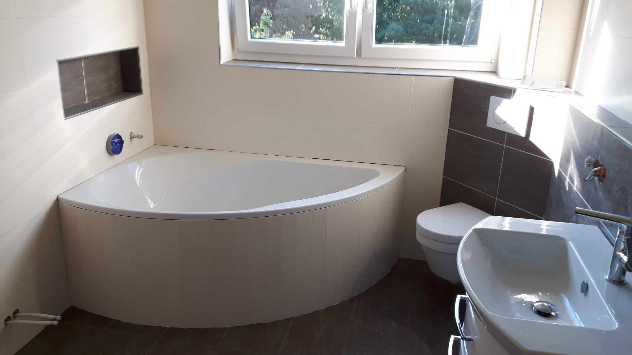 Jasna łazienka po renowacji z narożną wanną, umywalką na szafce i podwieszaną toaletą, wykończona białymi i szarymi płytkami z wnęką w ścianie.