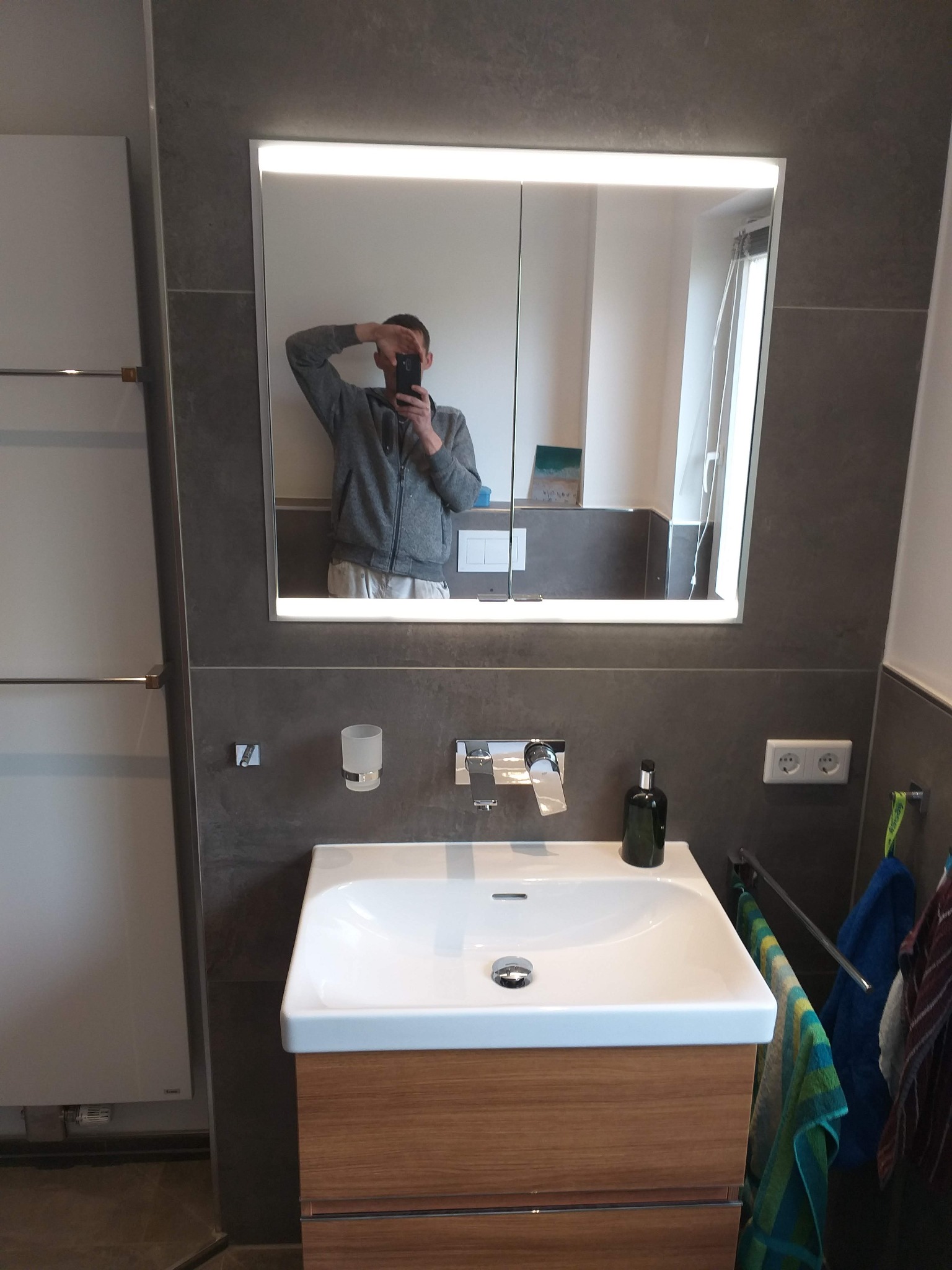 Nowoczesna łazienka z umywalką nablatową na drewnianej szafce, podświetlanym lustrem z odbijającym się fotografem i szarymi płytkami na ścianach.