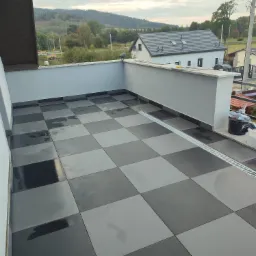 Szachownica z szarych i czarnych płytek na balkonie z odpływem liniowym, widok na góry w tle.