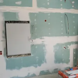 Ściana z płyt gipsowo-kartonowych w trakcie szpachlowania, widoczne puszki elektryczne, przewody, oraz zapas worków z zaprawą na podłodze.