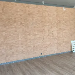 Ściana wykończona korkiem w odcieniach beżu i brązu, z widocznymi drobnymi prostokątnymi elementami, szara listwa przypodłogowa, panele podłogowe imitujące drewno, biała drabinka malarska w rogu...