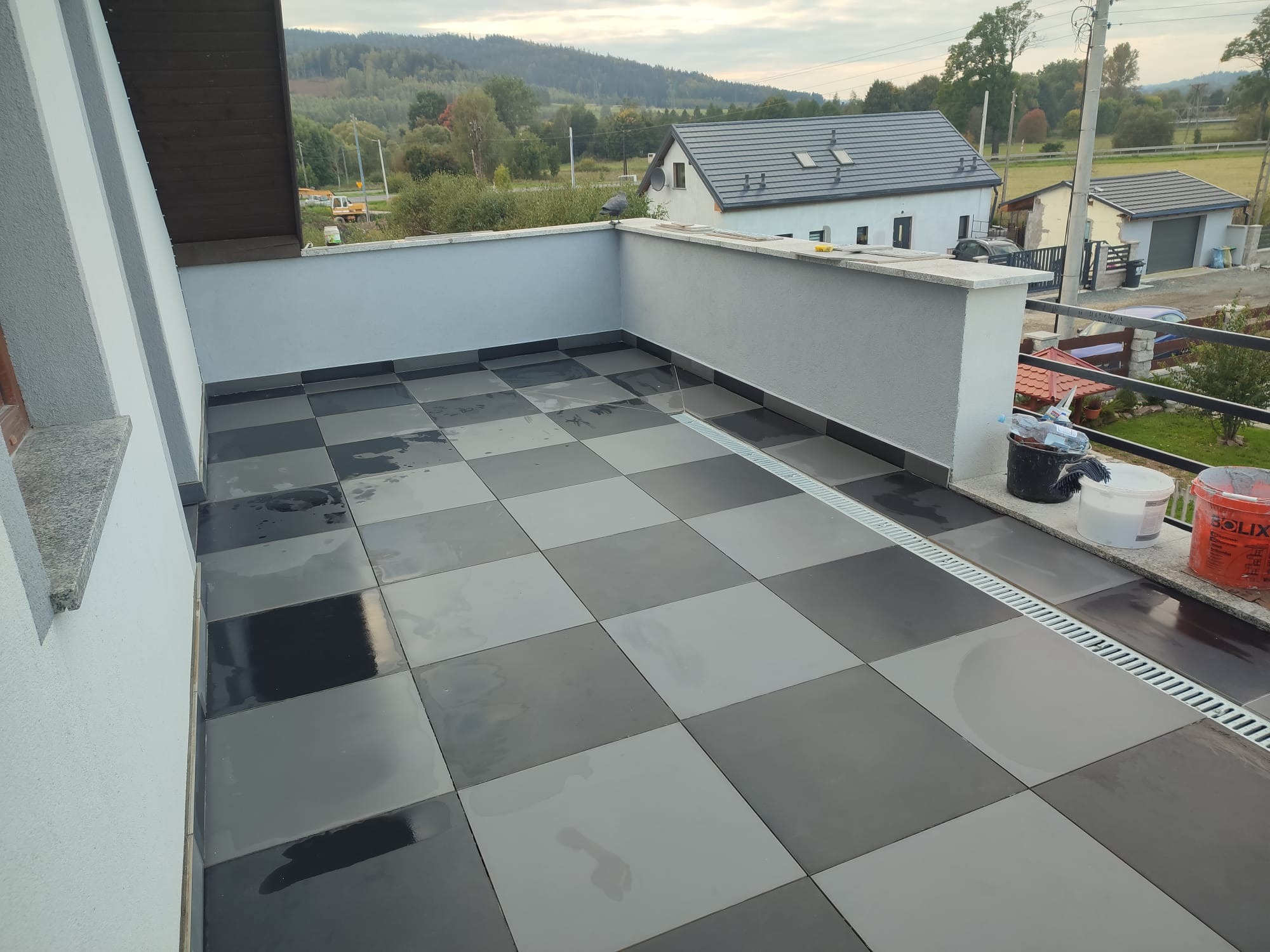 Szachownica z szarych i czarnych płytek na balkonie z odpływem liniowym, widok na góry w tle.